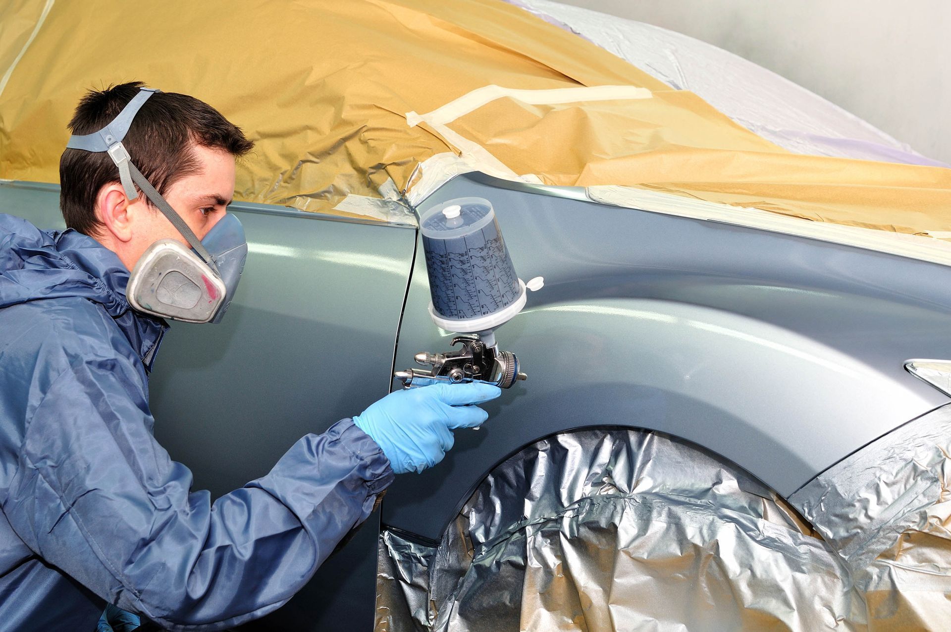 Painter spraying paint on a car fender in a repair shop, with masked and covered surfaces