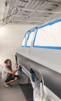Worker spraying a gray vehicle in a paint booth, masked off with blue tape and plastic.