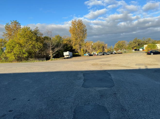 Empty parking lot under a cloudy sky, with trees and a few parked cars in the distance.