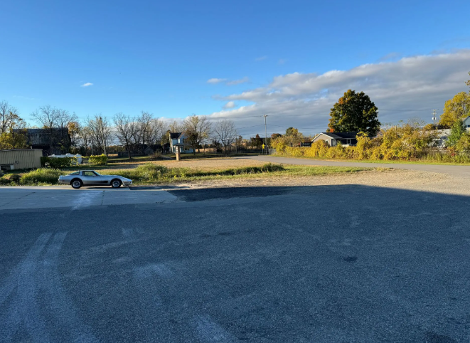 Open parking lot beside a grassy field under a blue sky, with a lone silver car and trees in autumn light.