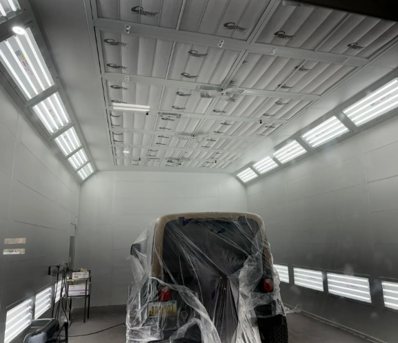 A car wrapped in plastic inside a bright white paint booth with overhead lights.