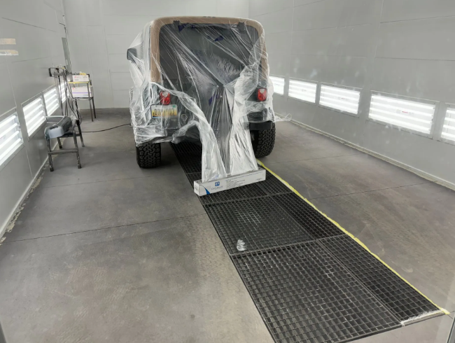 Black vehicle wrapped in plastic inside a spray booth, with a floor grate and yellow line nearby