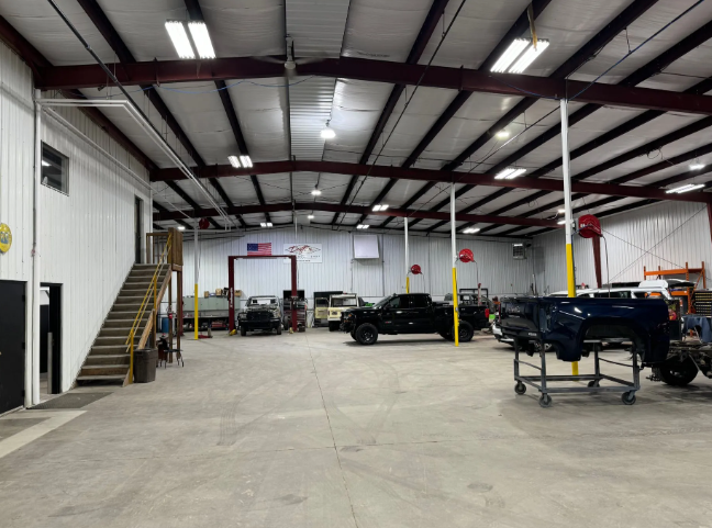 Large auto repair garage with trucks, lifts, and a staircase under a high metal ceiling