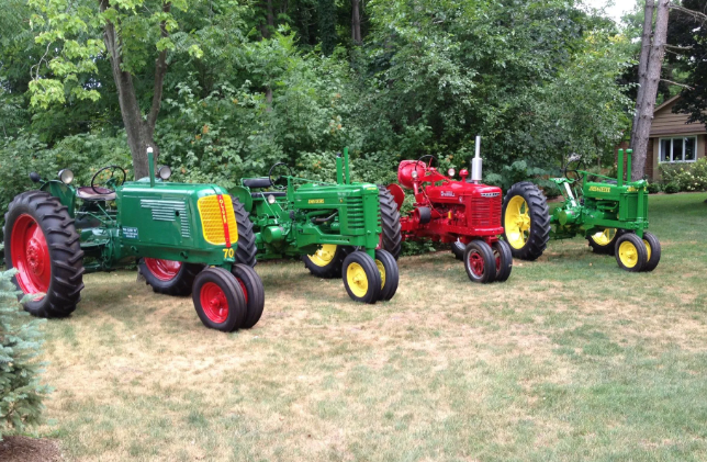 Three colorful vintage tractors lined up on grass with trees in the background
