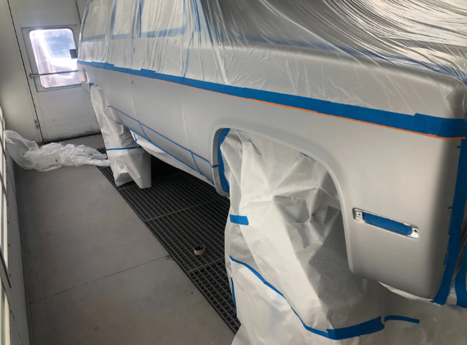 Car in a paint booth, covered in plastic masking and blue tape, with wheel and window protected.