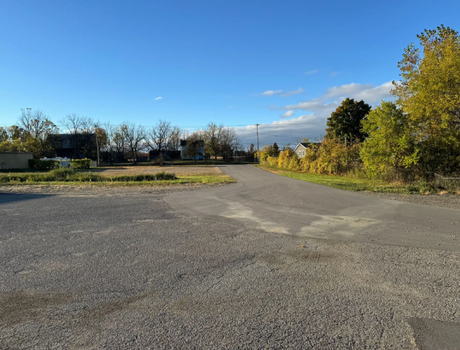 Empty paved lot beside a road under a blue sky, with trees and low buildings in the distance