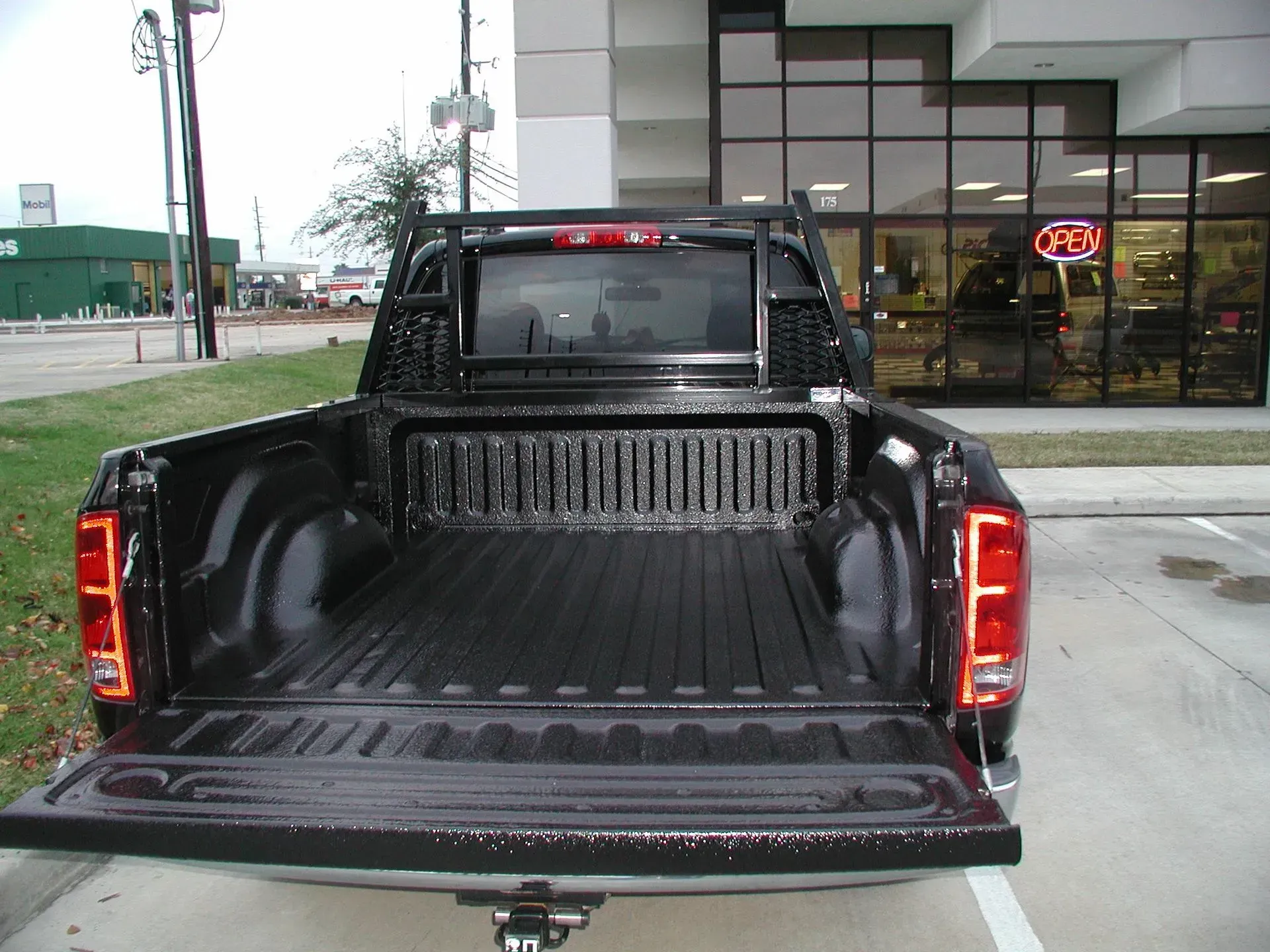 Black pickup truck bed with open tailgate, spray-in bed liner, and head rack, parked in front of a store.