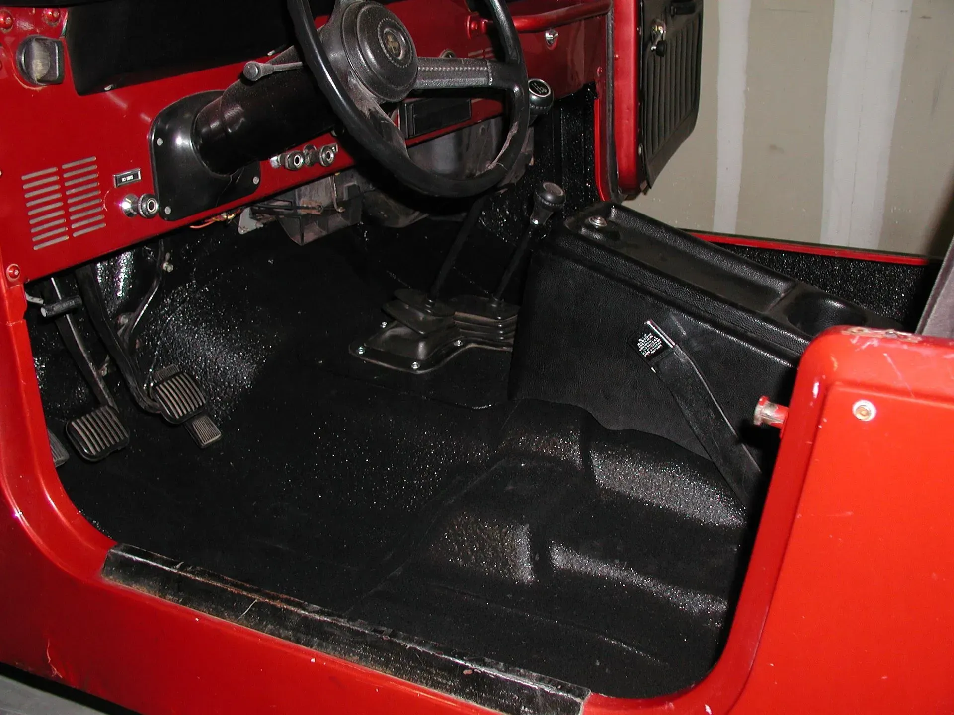 Interior of a red Jeep with black textured flooring, steering wheel, and pedals.