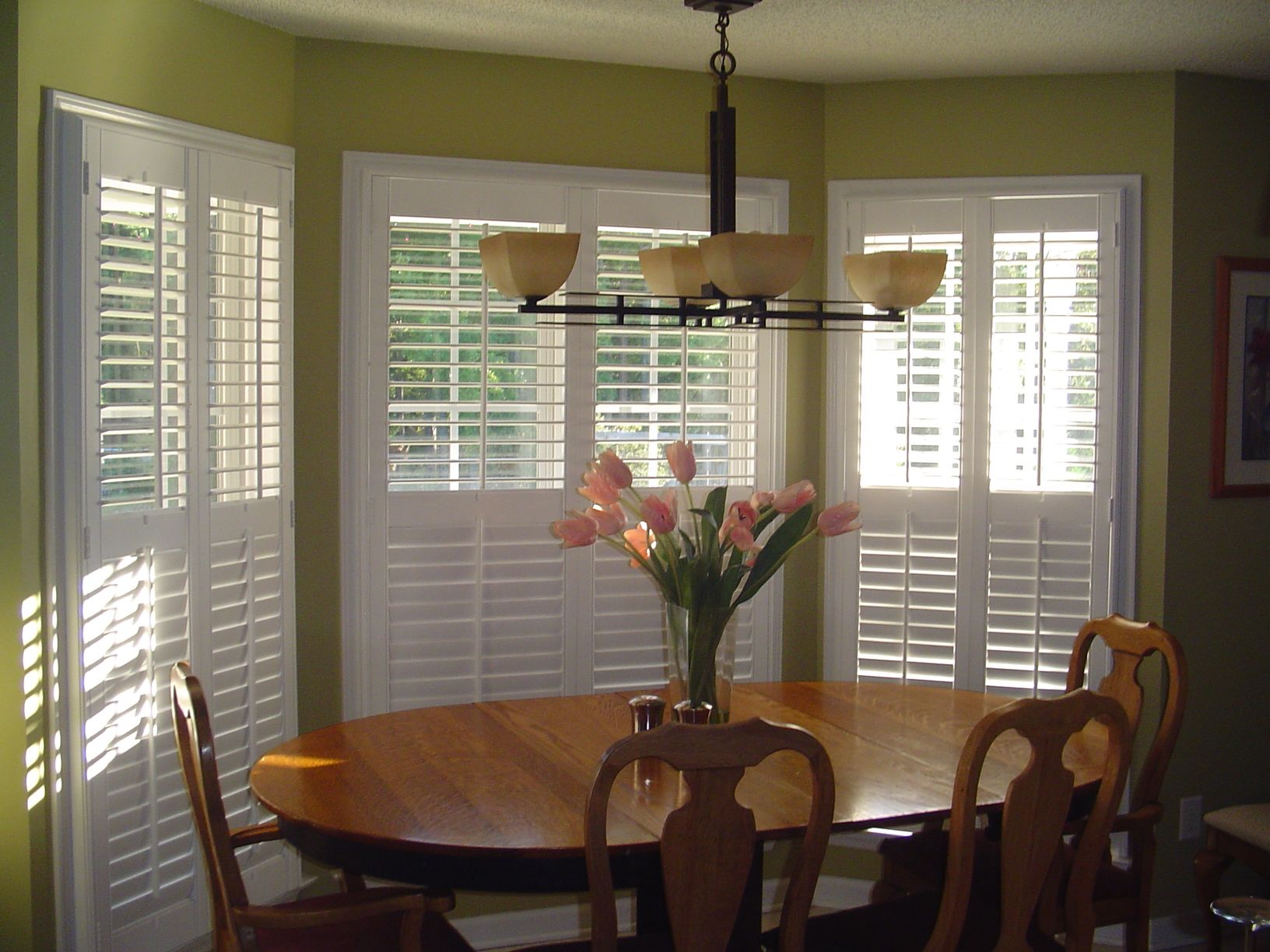 Dining room with oval table, chairs, and window shutters. Flowers on the table. Sunlight.