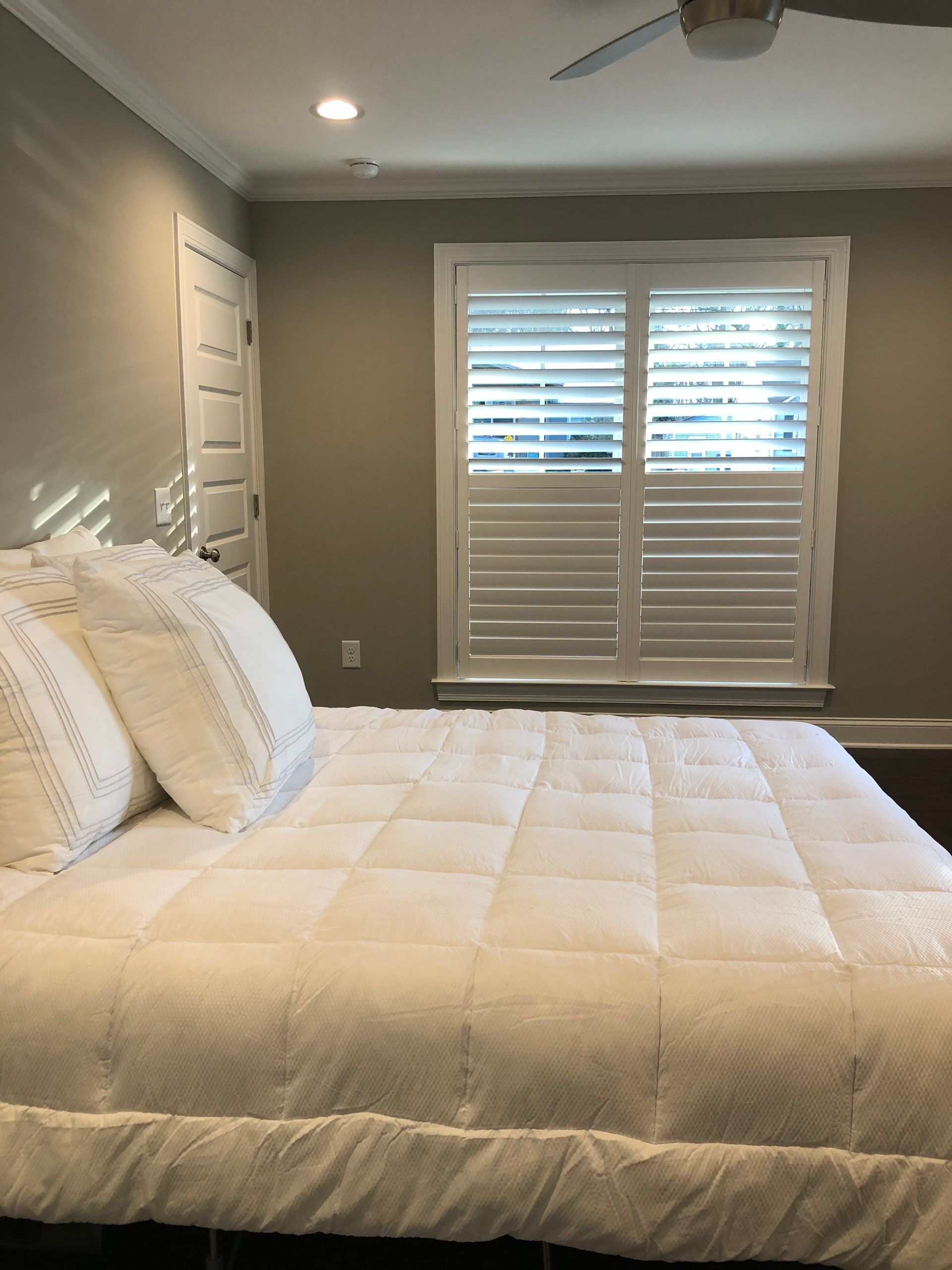 White bed with fluffy bedding, a window with shutters, and a door in a beige-walled bedroom.