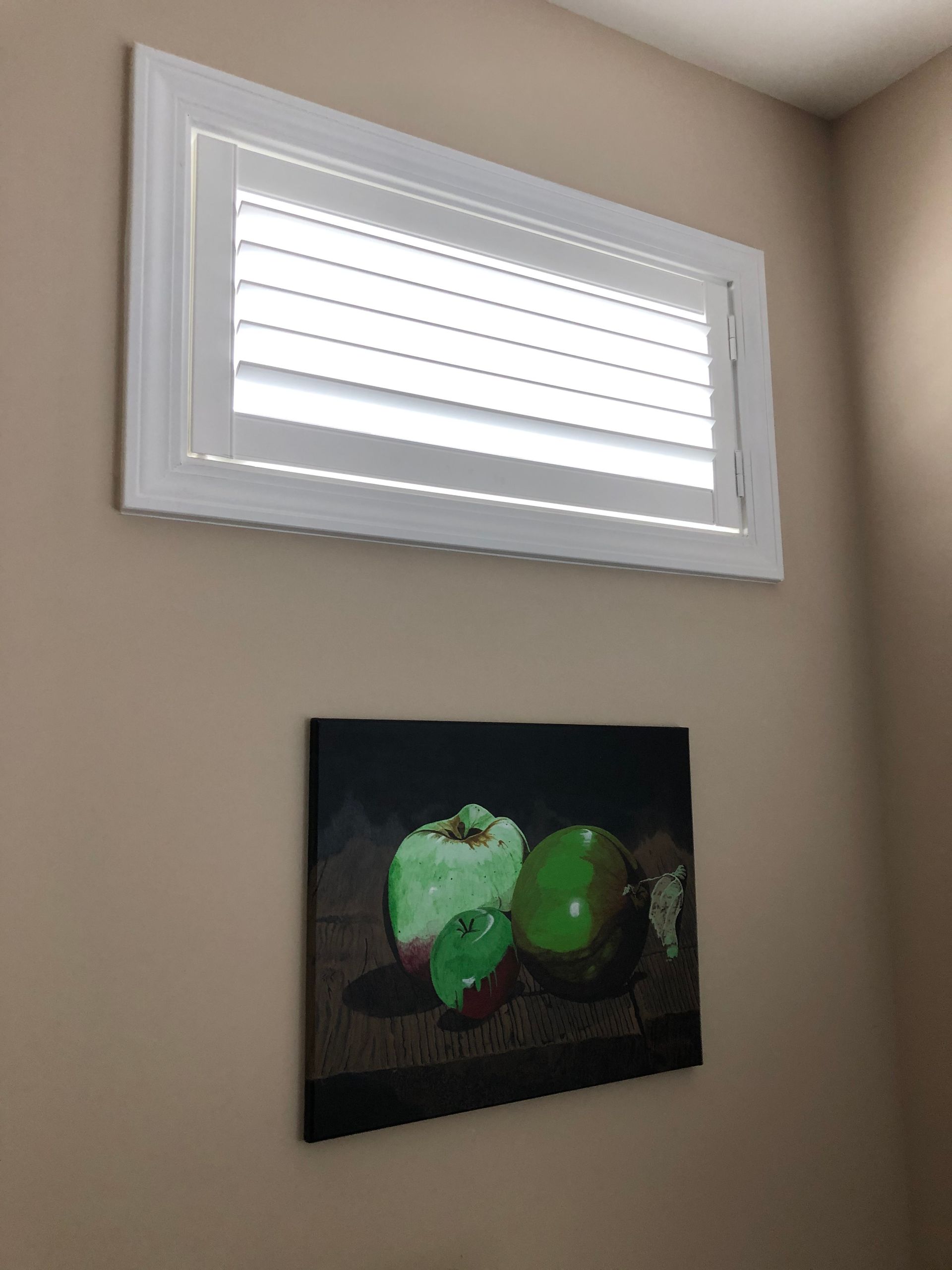 White-framed window with closed blinds above a painting of green apples on a wall.