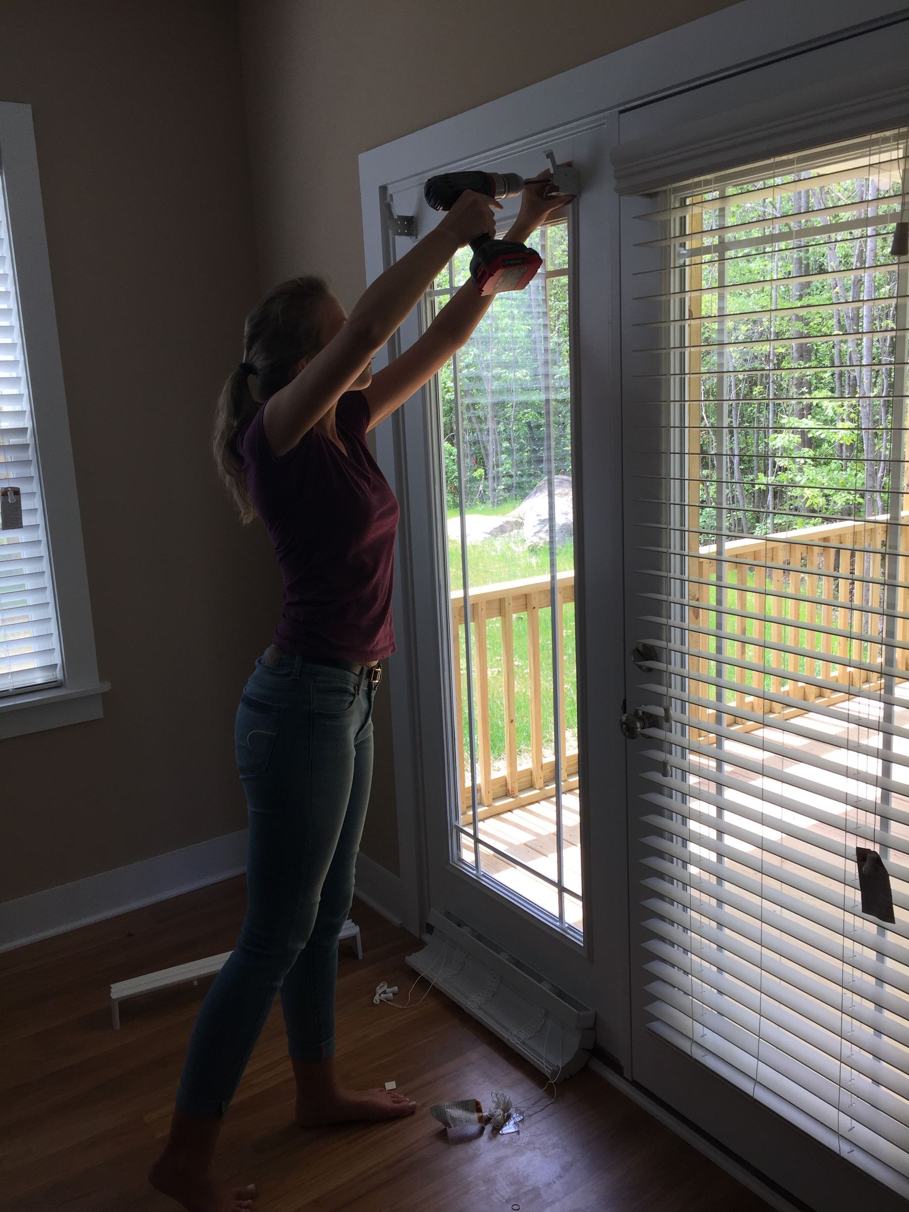 Woman using a drill to install blinds on a sliding glass door. Interior shot with wooden deck visible outside.