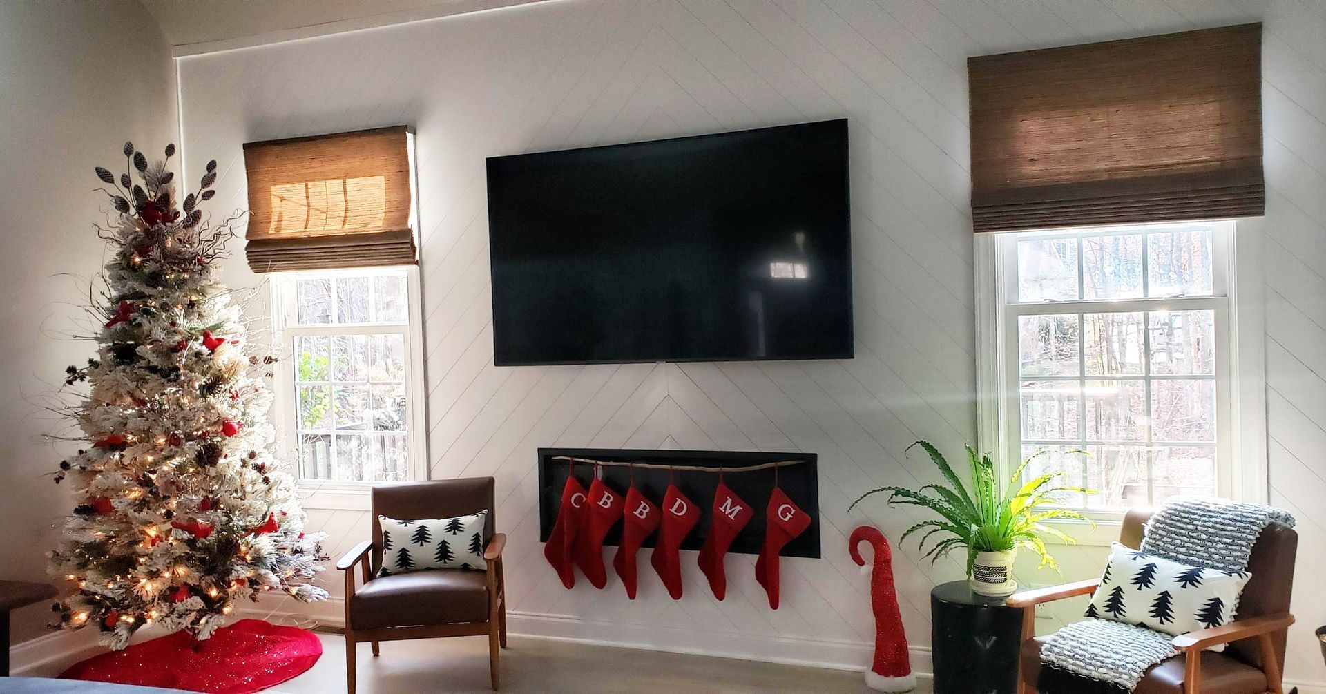 Living room with Christmas tree, TV above fireplace, red stockings. Two chairs by windows.