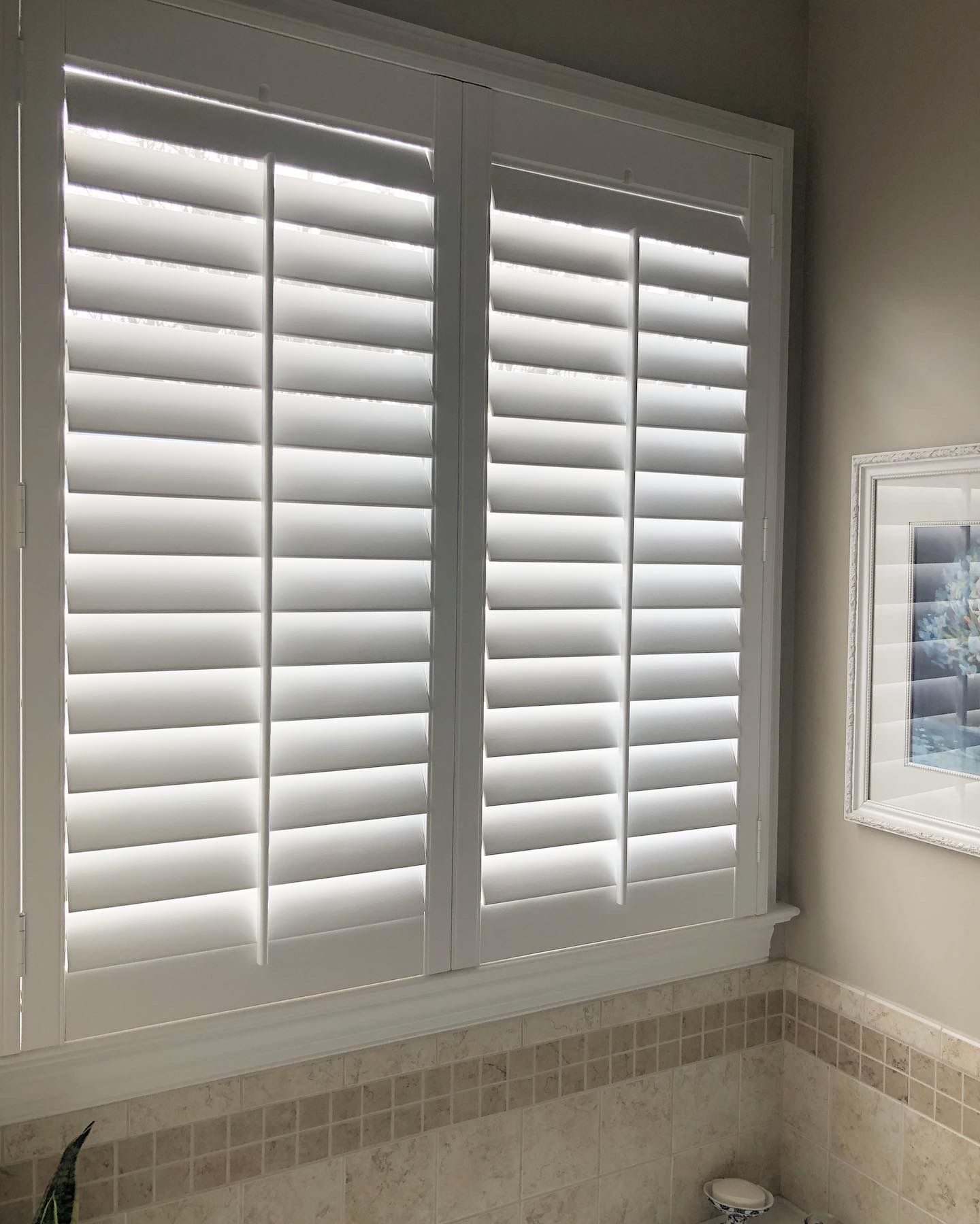 White window shutters inside a bathroom with beige tile and a framed picture.