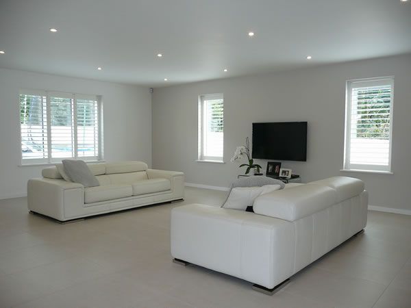 Modern living room with white leather sofas, a mounted TV, and shuttered windows.