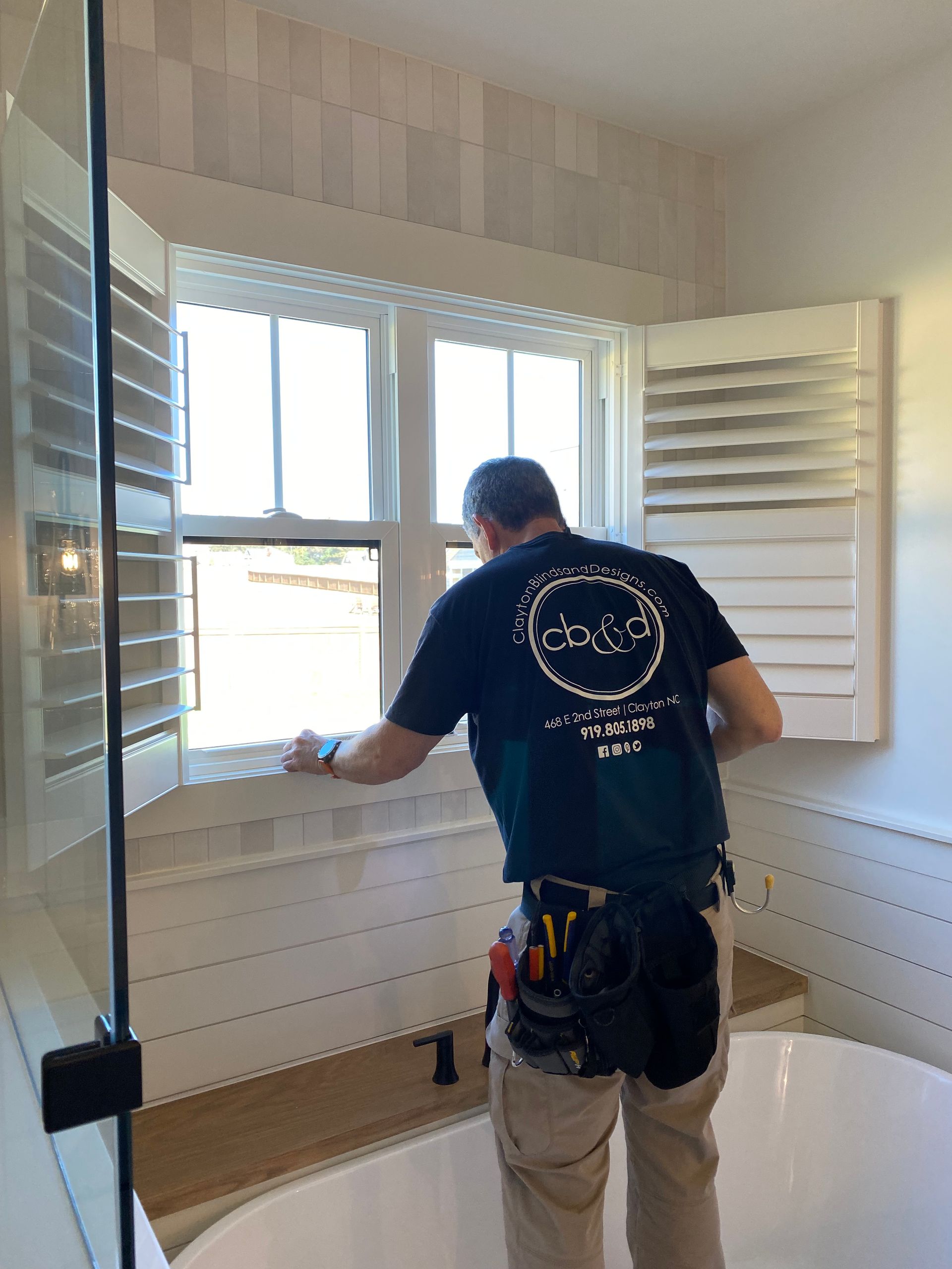 Man installing white shutters in a bathroom with a tub and window.
