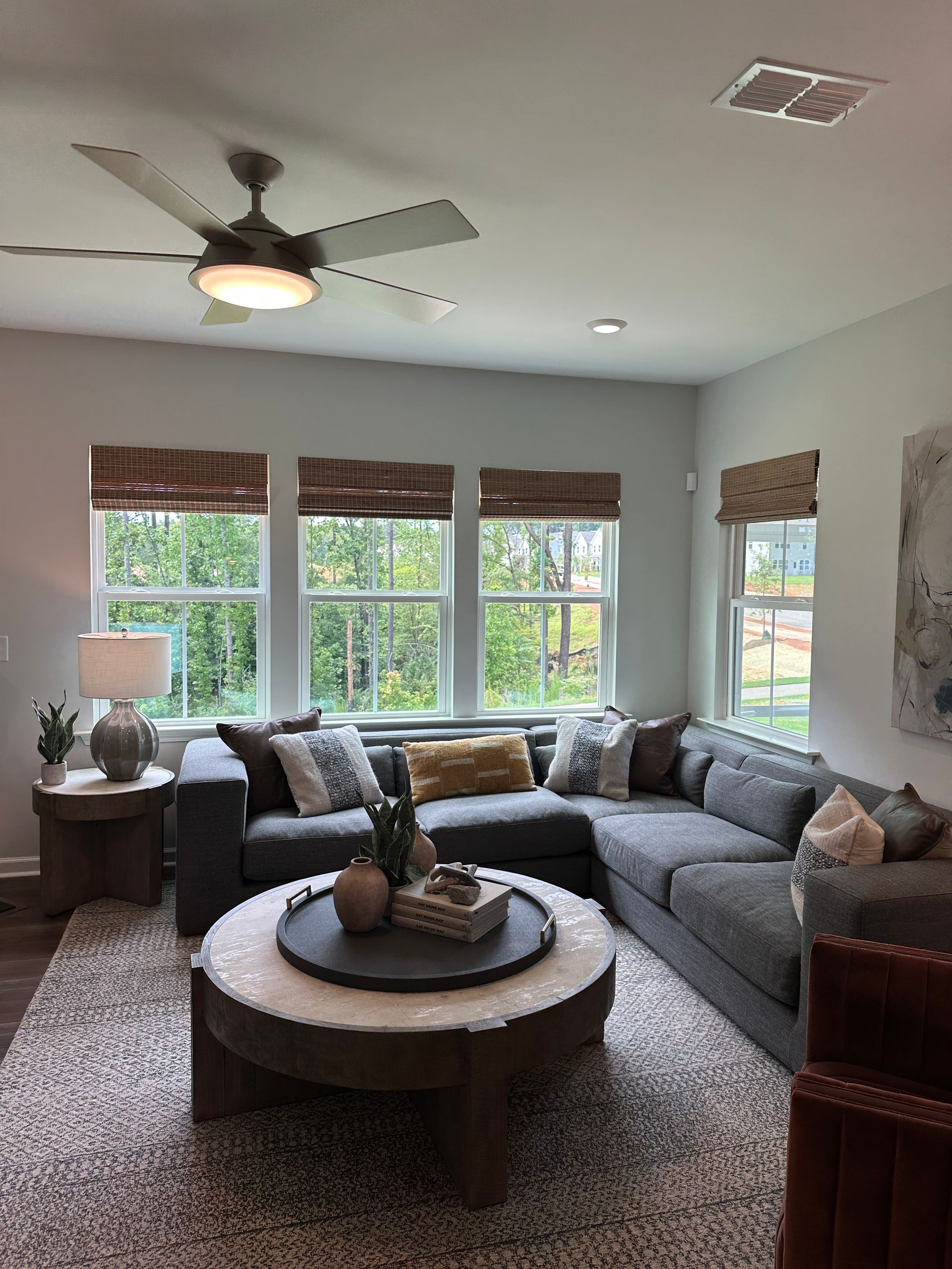 Living room with gray sectional sofa, round coffee table, and three windows with natural shades.