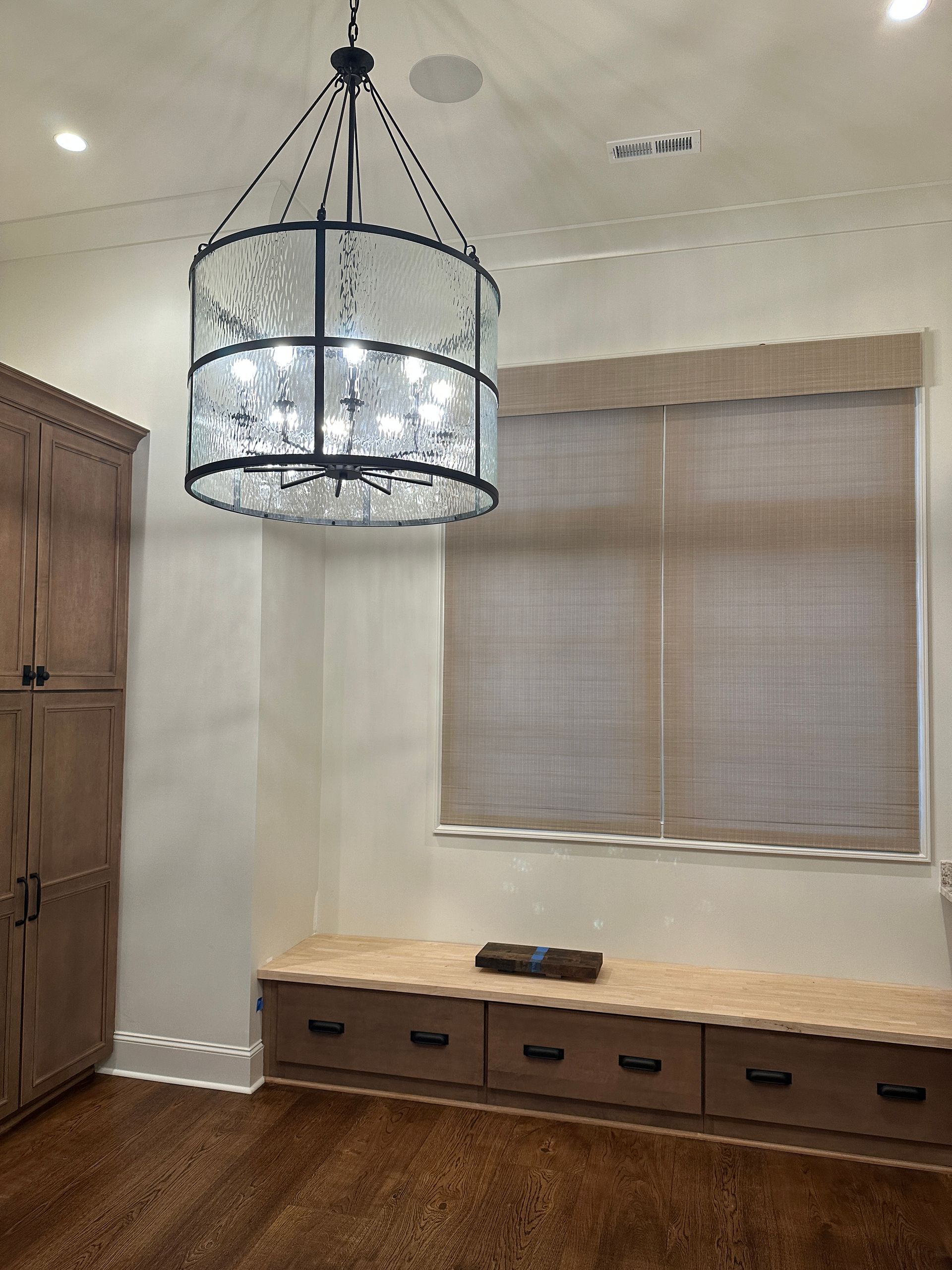 A room with a wooden cabinet, a bench, and a large chandelier with a window that has blinds.
