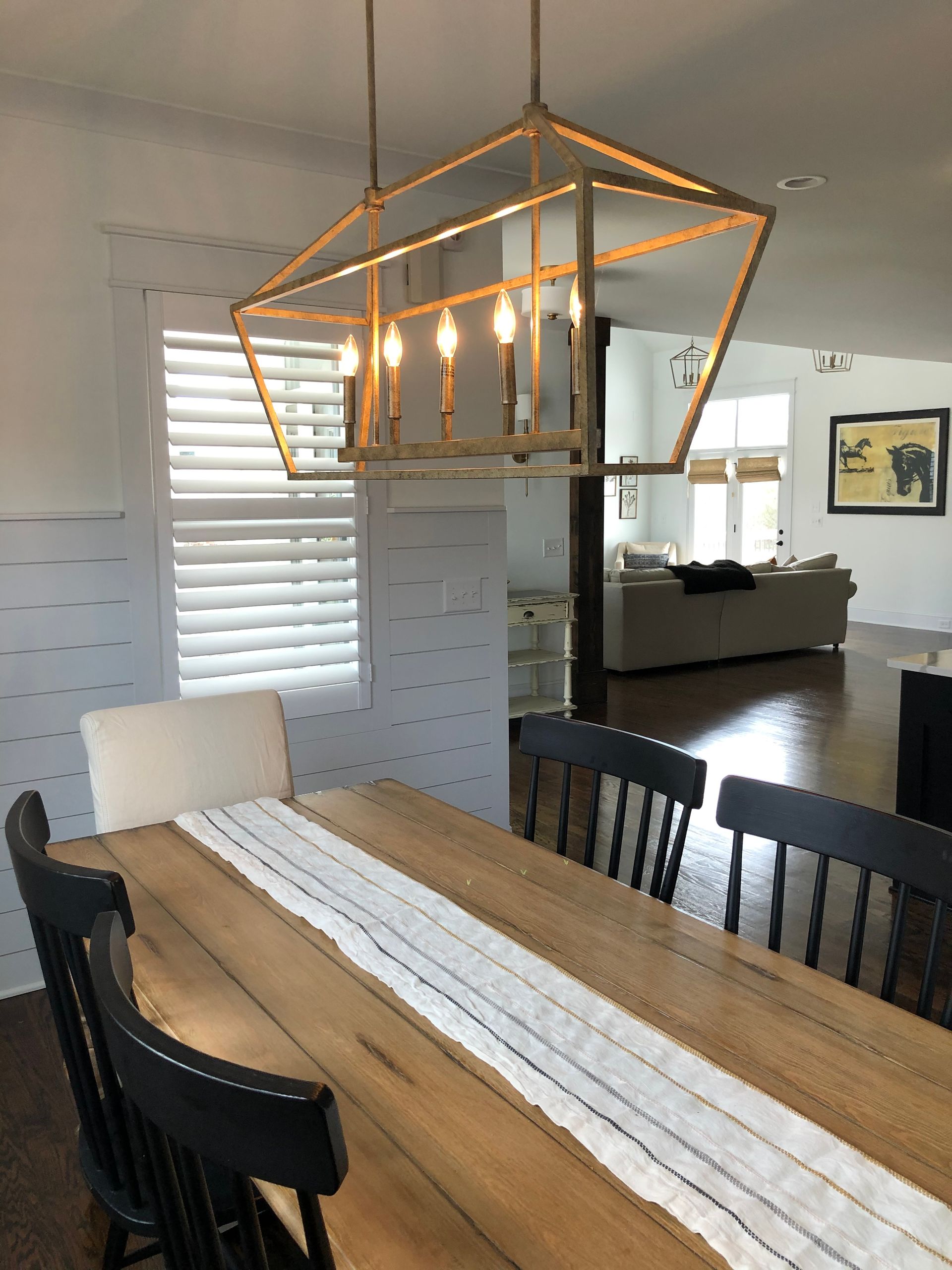 Dining room with wooden table, black chairs, and geometric light fixture. White shutters in the background.