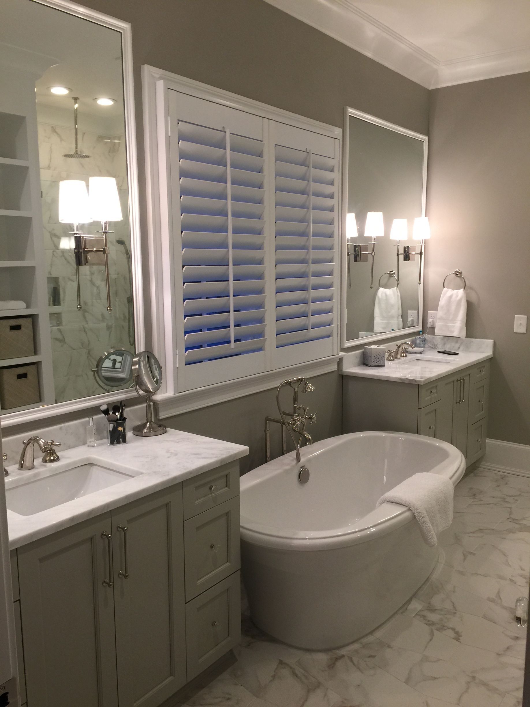 Elegant bathroom with a free-standing tub, two vanities, large mirrors, and shuttered window. Gray and white color scheme.