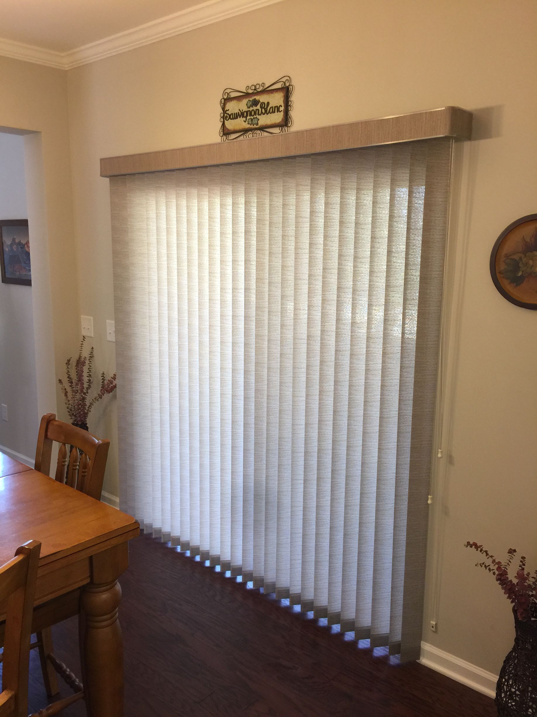 Beige vertical blinds cover a sliding glass door in a dining room. Wooden table and vase of flowers in view.