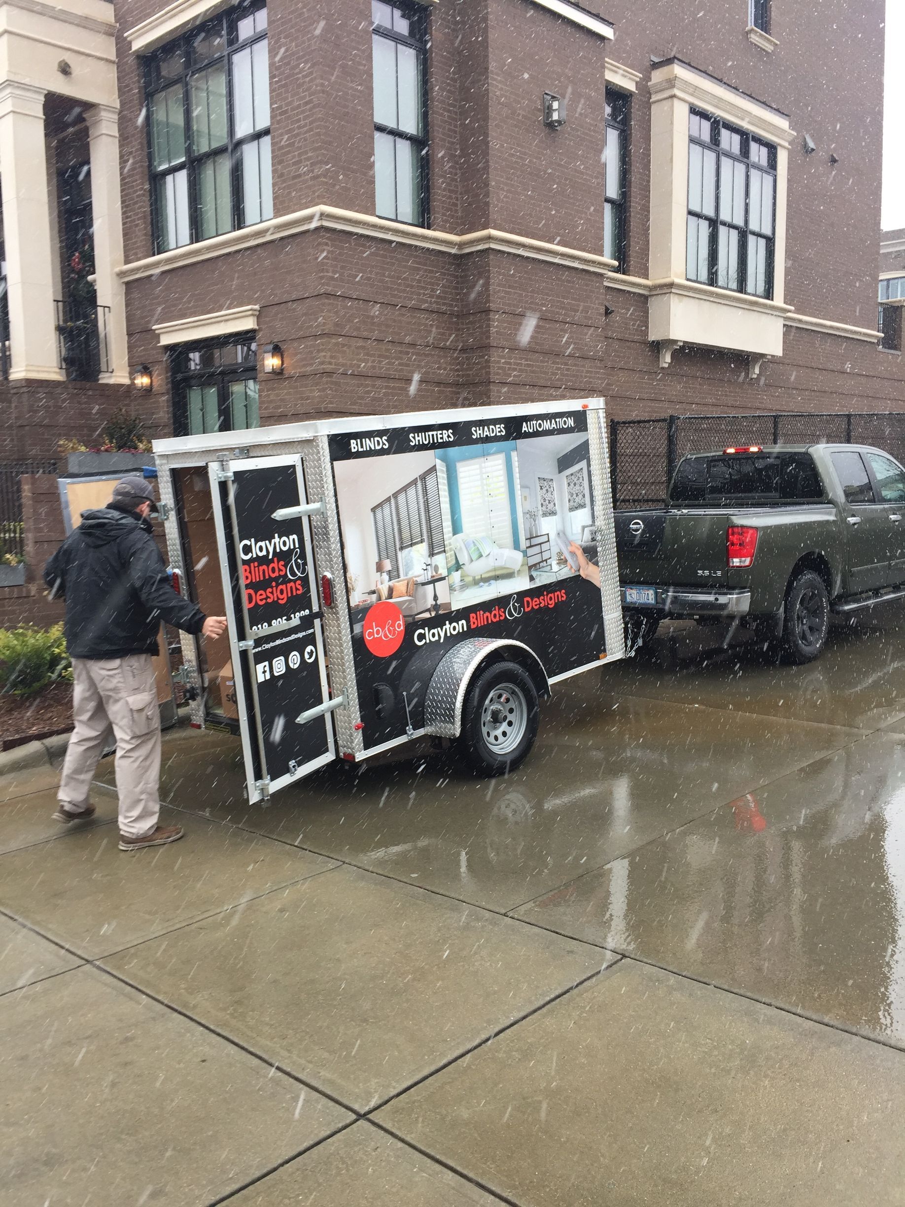 Man opening trailer door hitched to truck in snowy conditions near building.