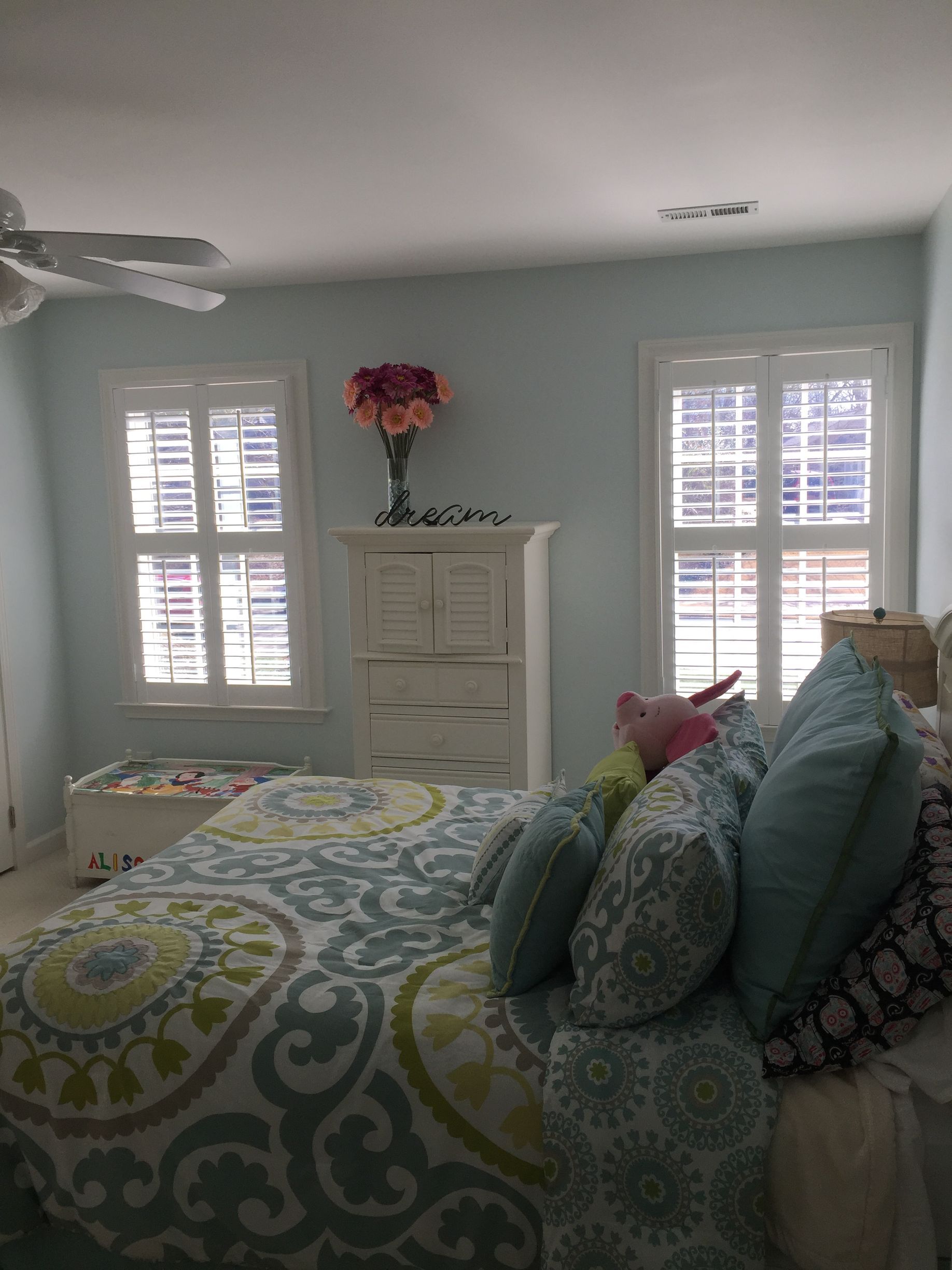 Bedroom with blue walls, two windows with shutters, white dresser with flowers, patterned bedding, and pillows.