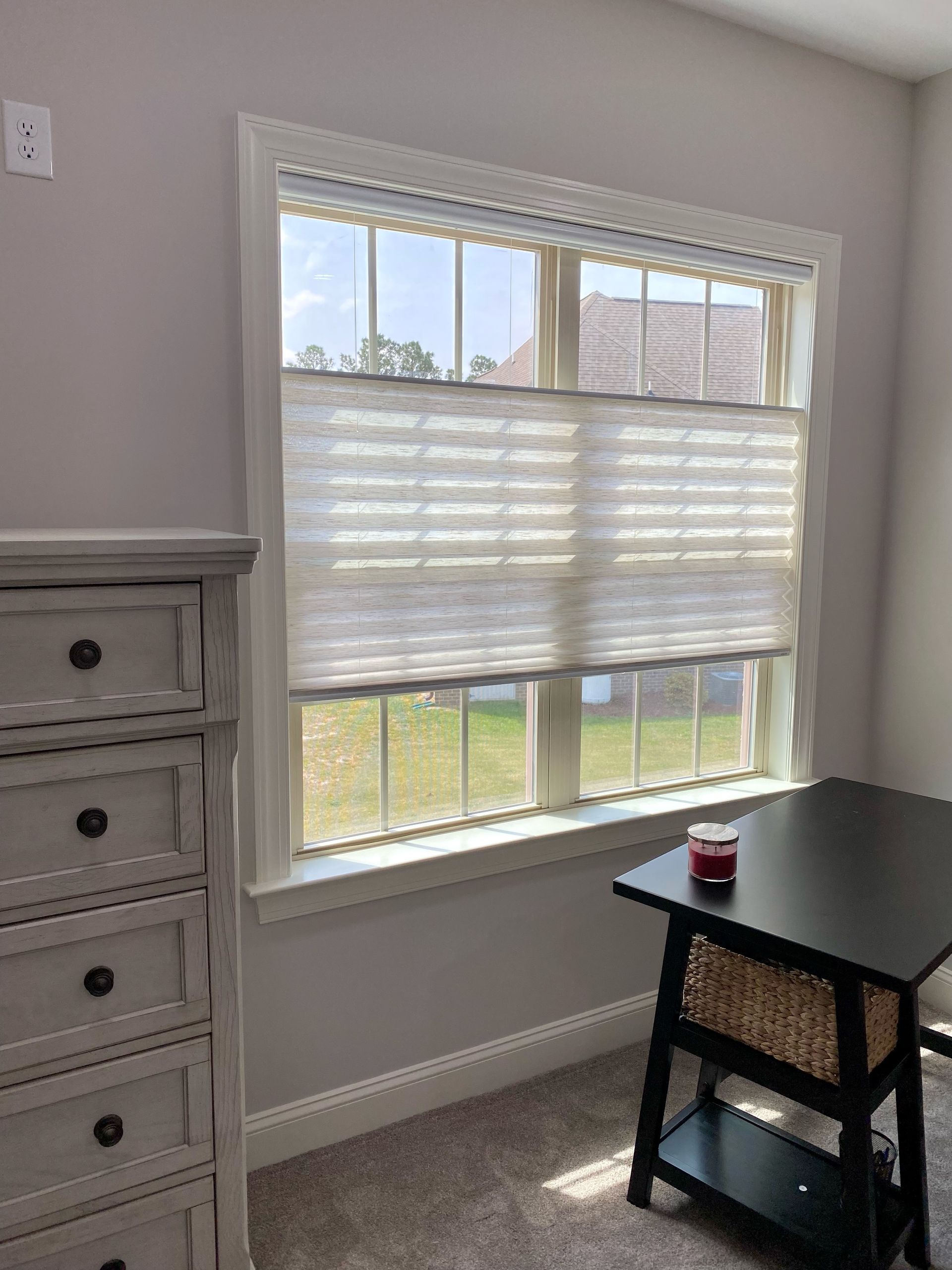 A window with white cellular shades, overlooking a backyard. A dresser and small desk are nearby.