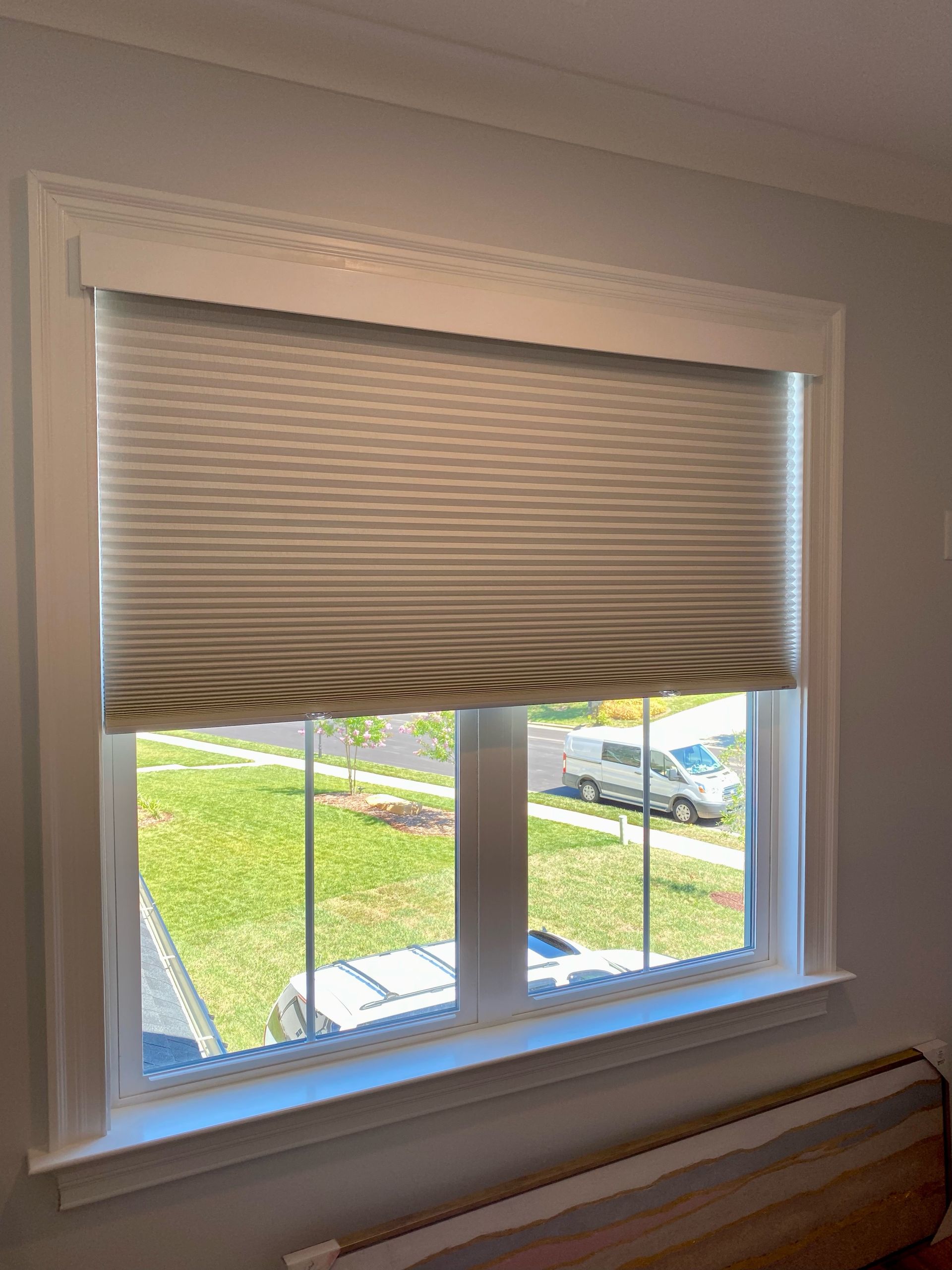Window with closed pleated blinds, white frame, view of outside with green grass and a vehicle.