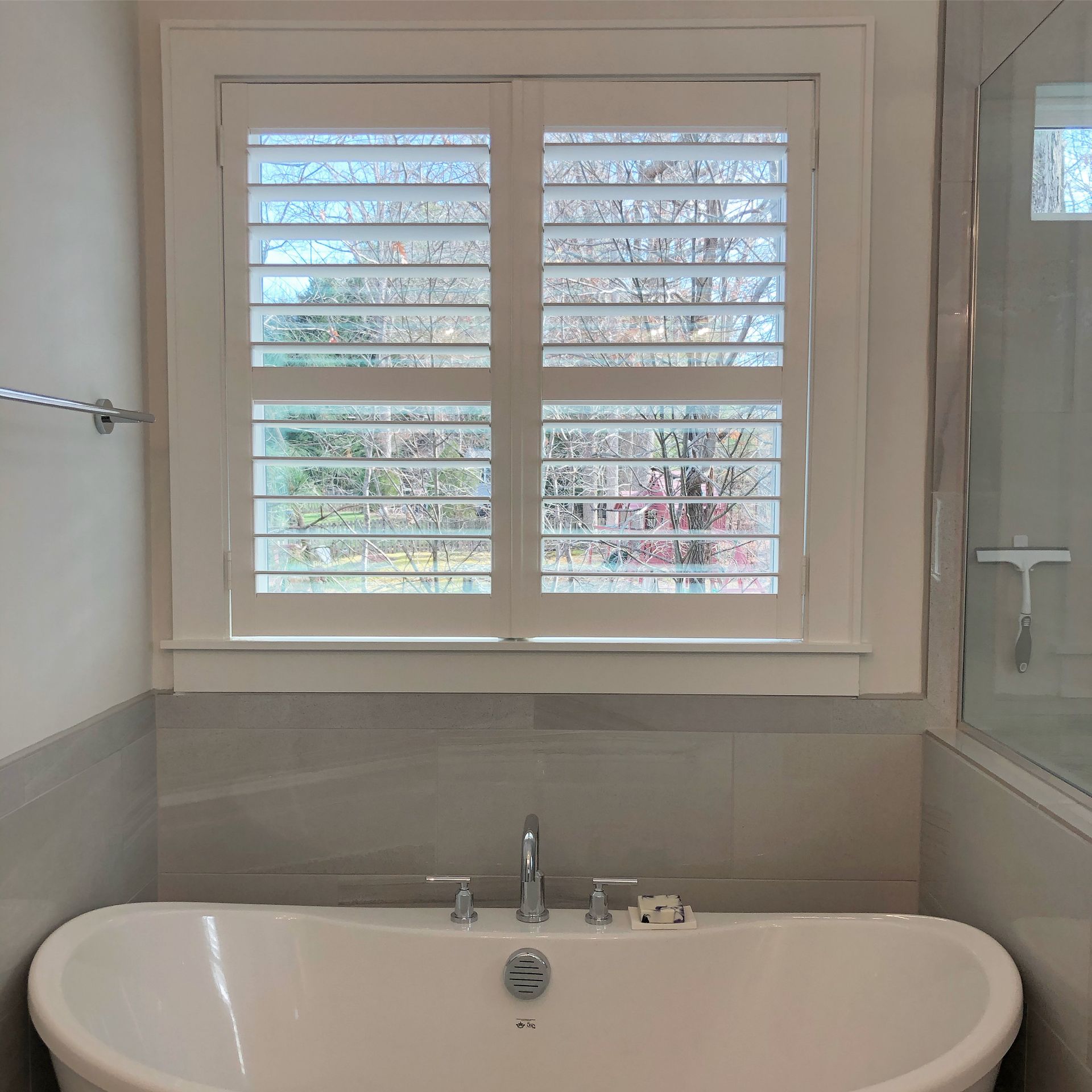 Bathroom with white shutters over a window, above a white tub with silver fixtures.