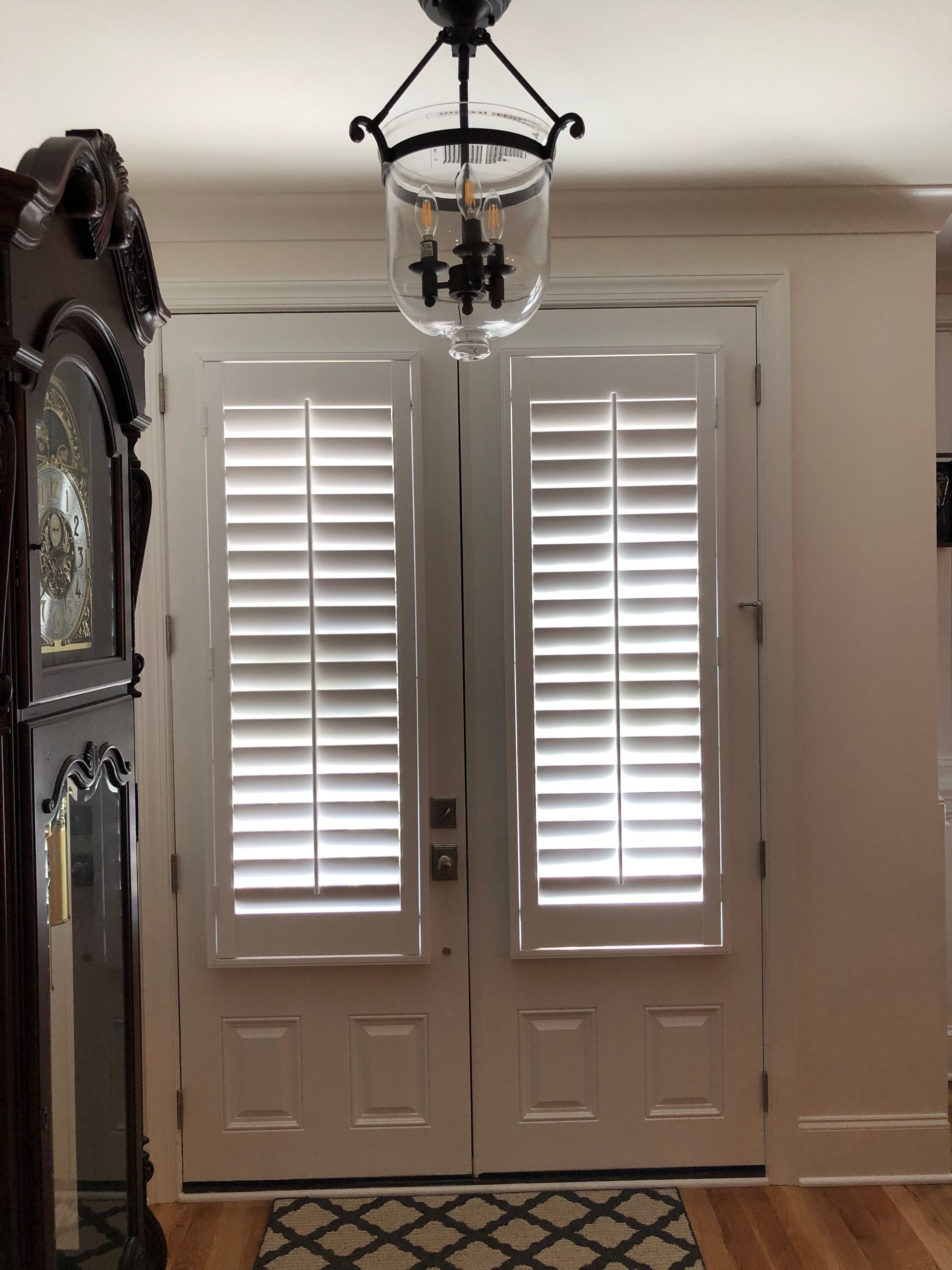 Double white doors with shutters, topped by a glass chandelier. A dark grandfather clock is on the left.