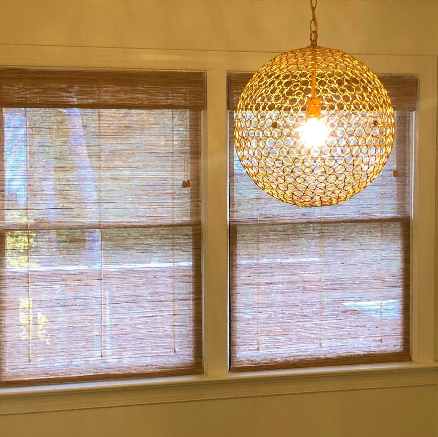 Two windows with brown woven shades and a gold, orb-shaped chandelier.