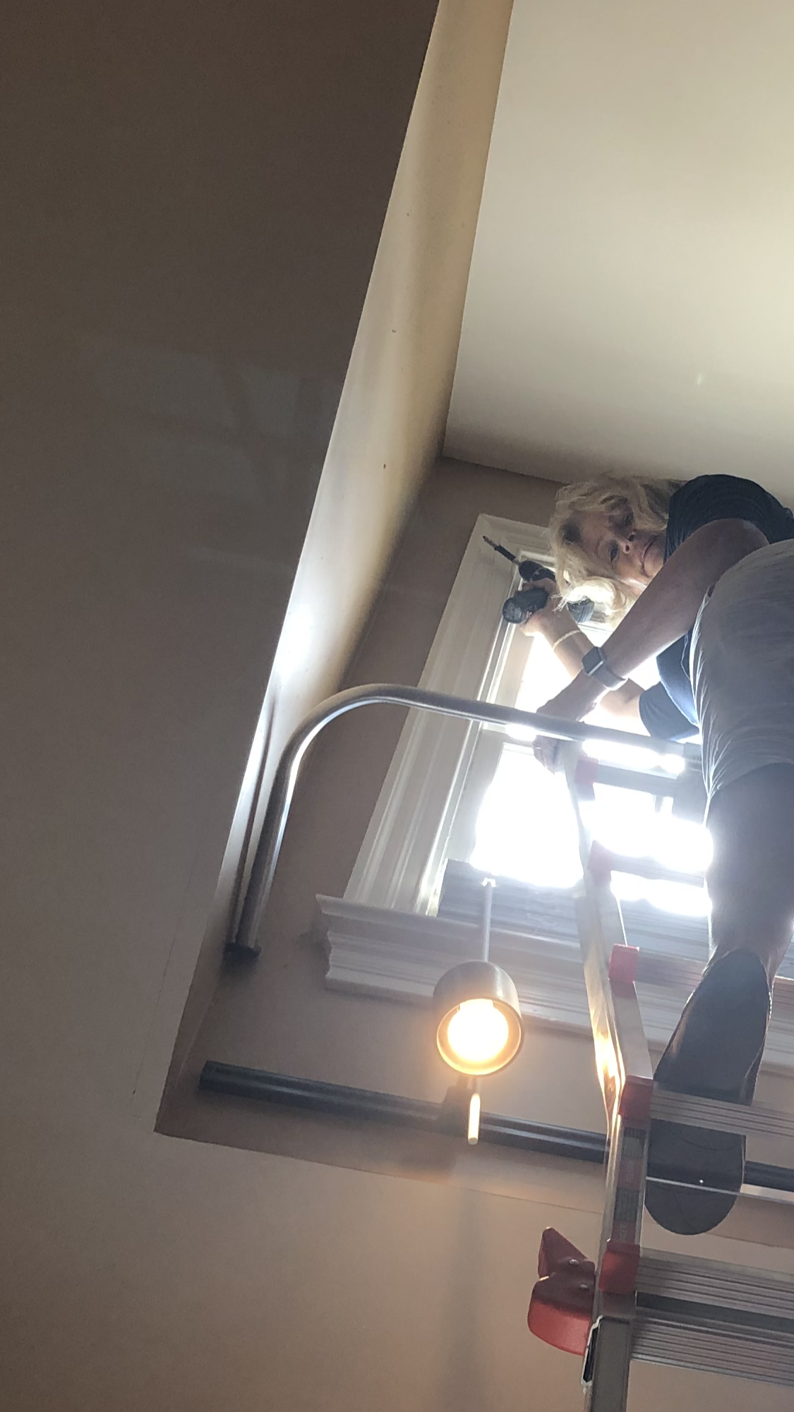 Person using a power tool, standing on a step ladder near a window. Sunlight streams through the window.