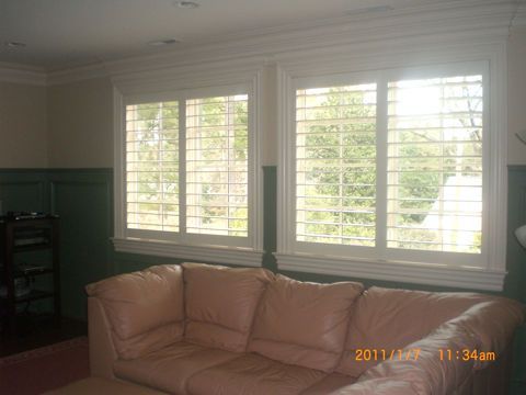 Living room with white shuttered windows, tan couch, and green paneling. Sunlight streams through the windows.