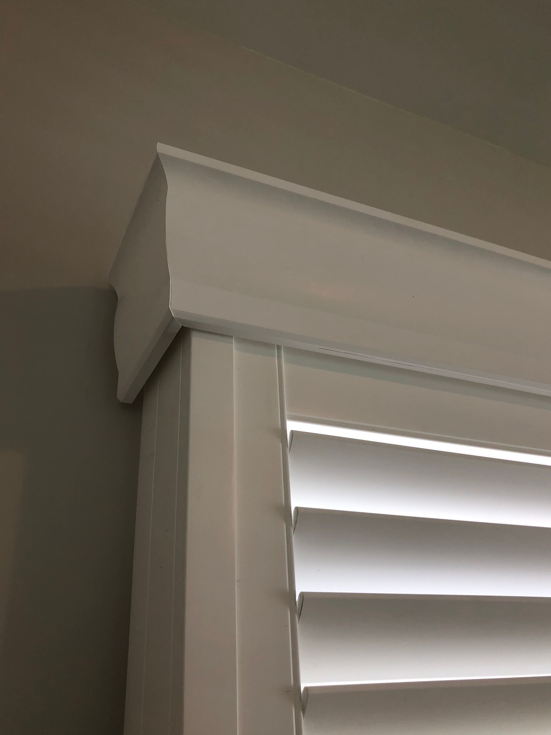 White shutter blinds with top valance attached to a white window frame, against a white wall.