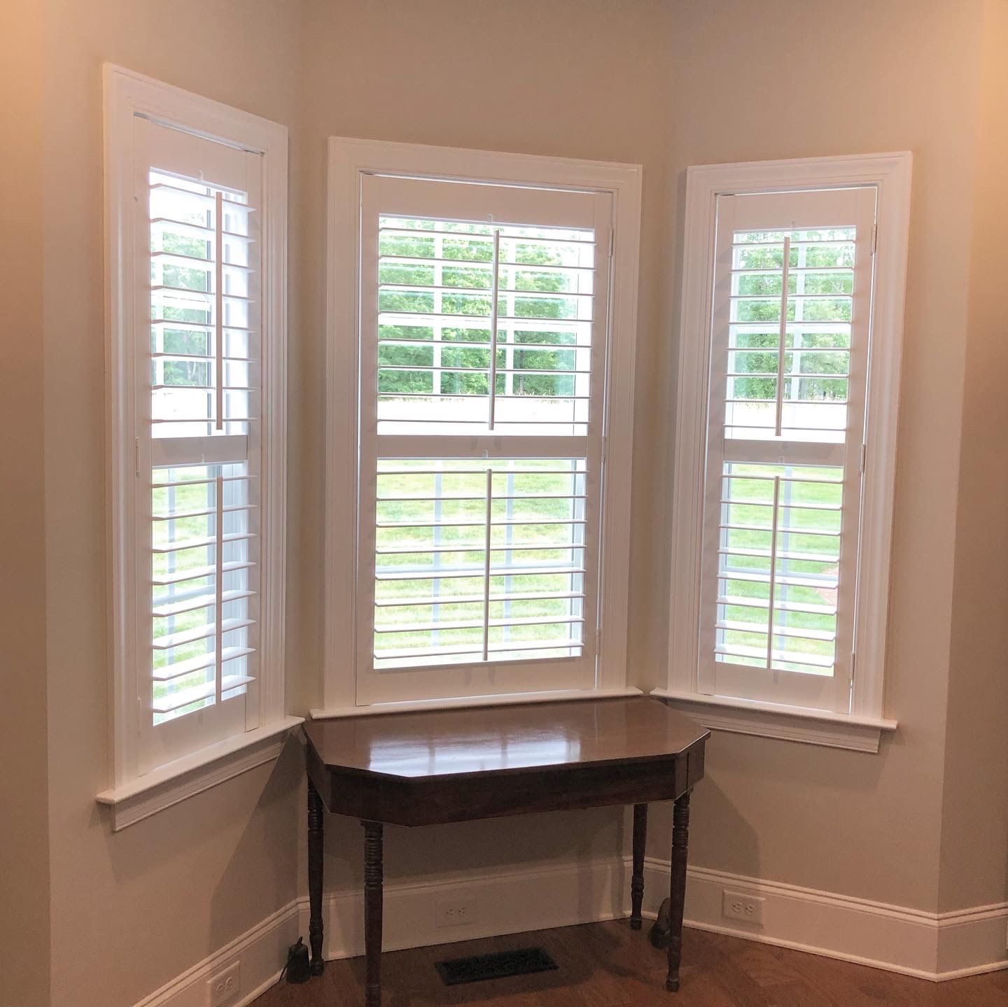 Bay window with white shutters, a wooden table, and beige walls.