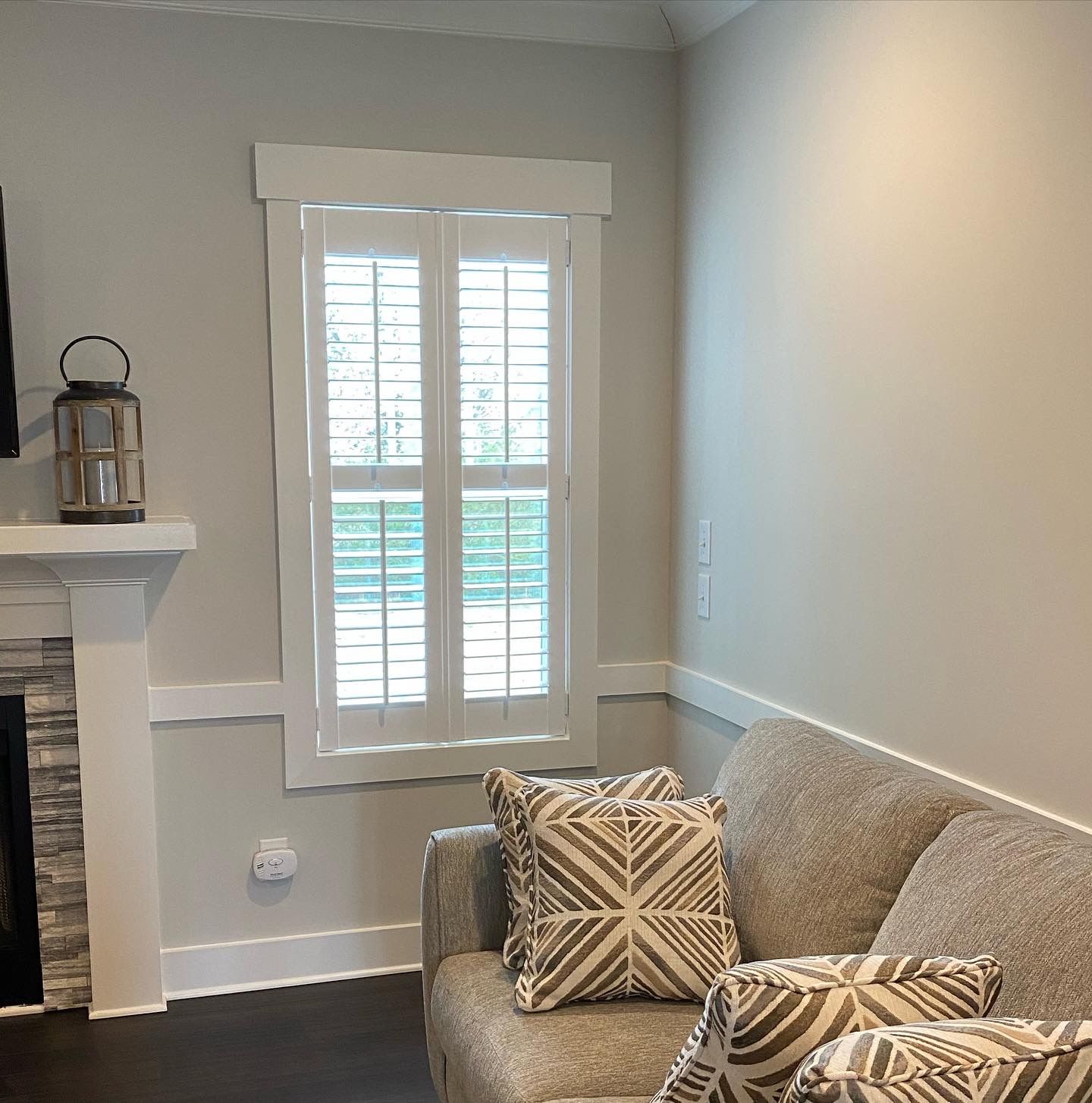 Living room corner with a window, white shutters, and a gray couch with patterned pillows.