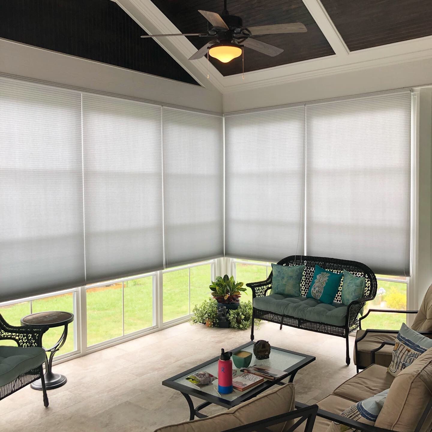Sunroom with gray blinds, black ceiling, wicker furniture, and potted plants.