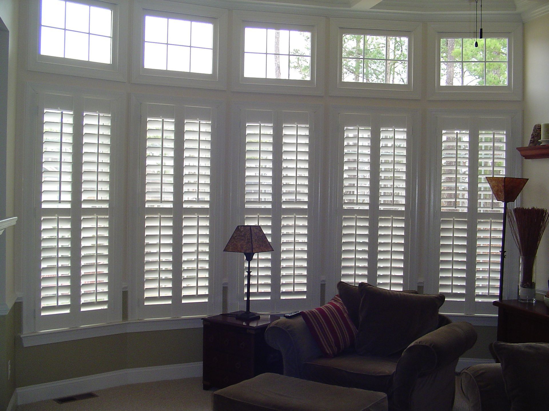 Bay window with white shutters, upper windows, living room with furniture, and a lamp.