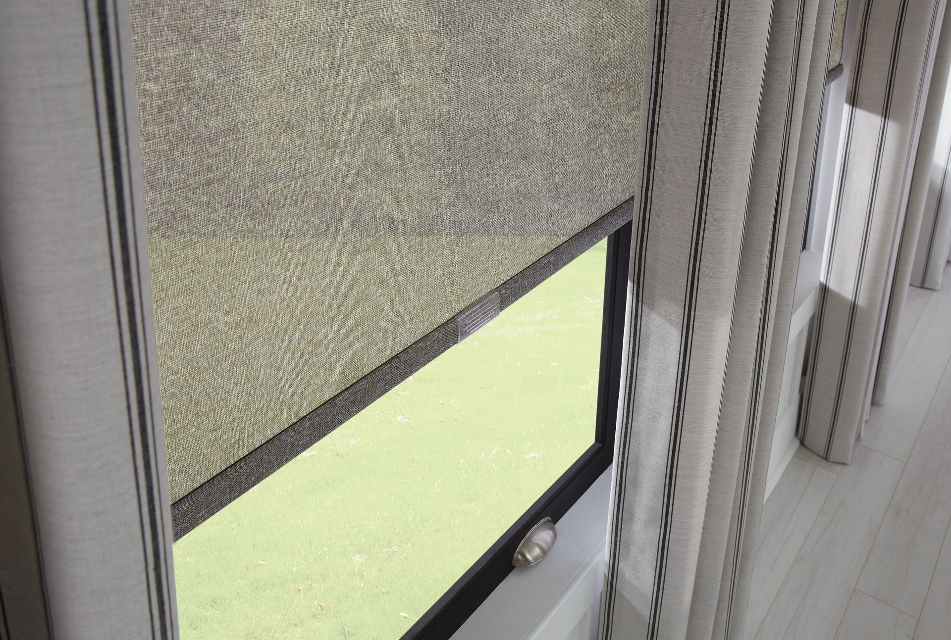 Beige roller blind partially covering a window, with striped curtains on the right.