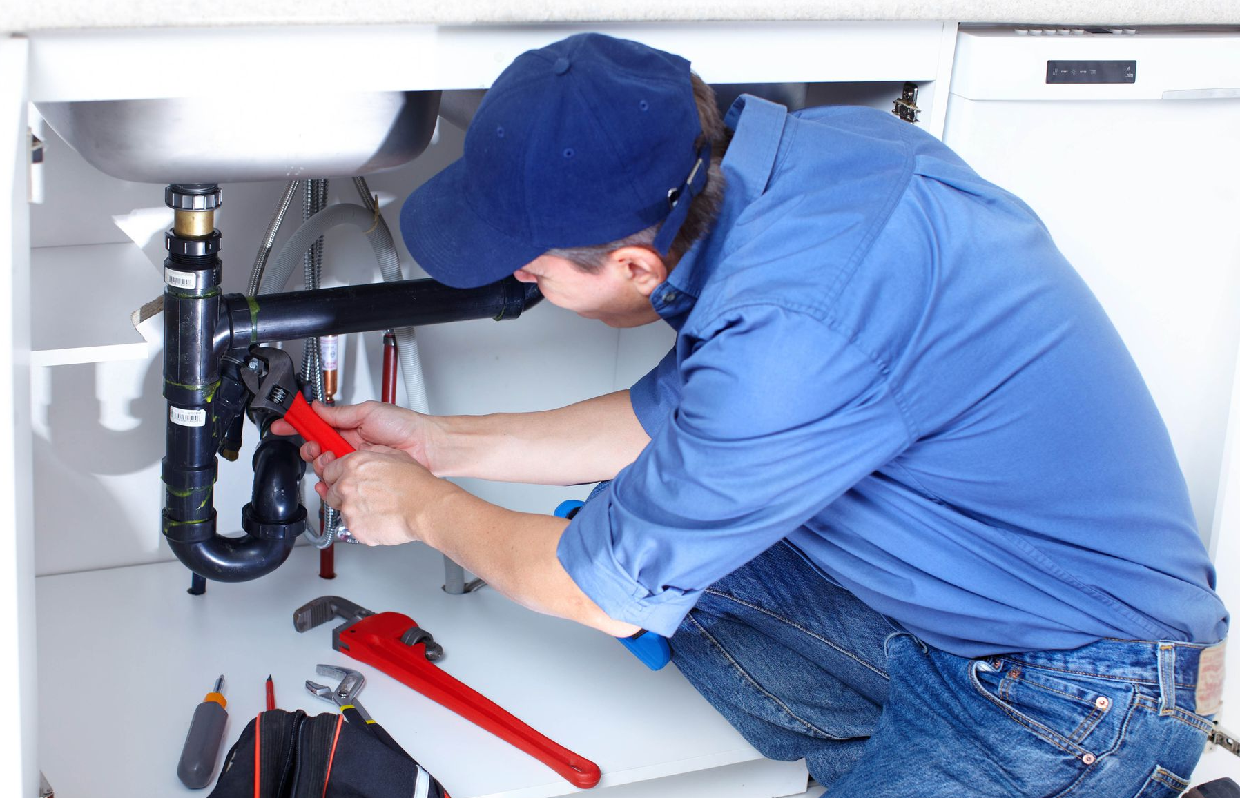 Plumber in blue shirt and hat working under a kitchen sink, using red wrench on pipes.
