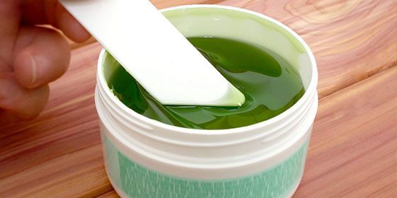 A hand holding a white spatula dips into a jar of green gel.
