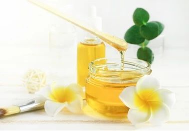 Honey dripping from a wooden stick into a jar, flowers, and green leaves in a light setting.