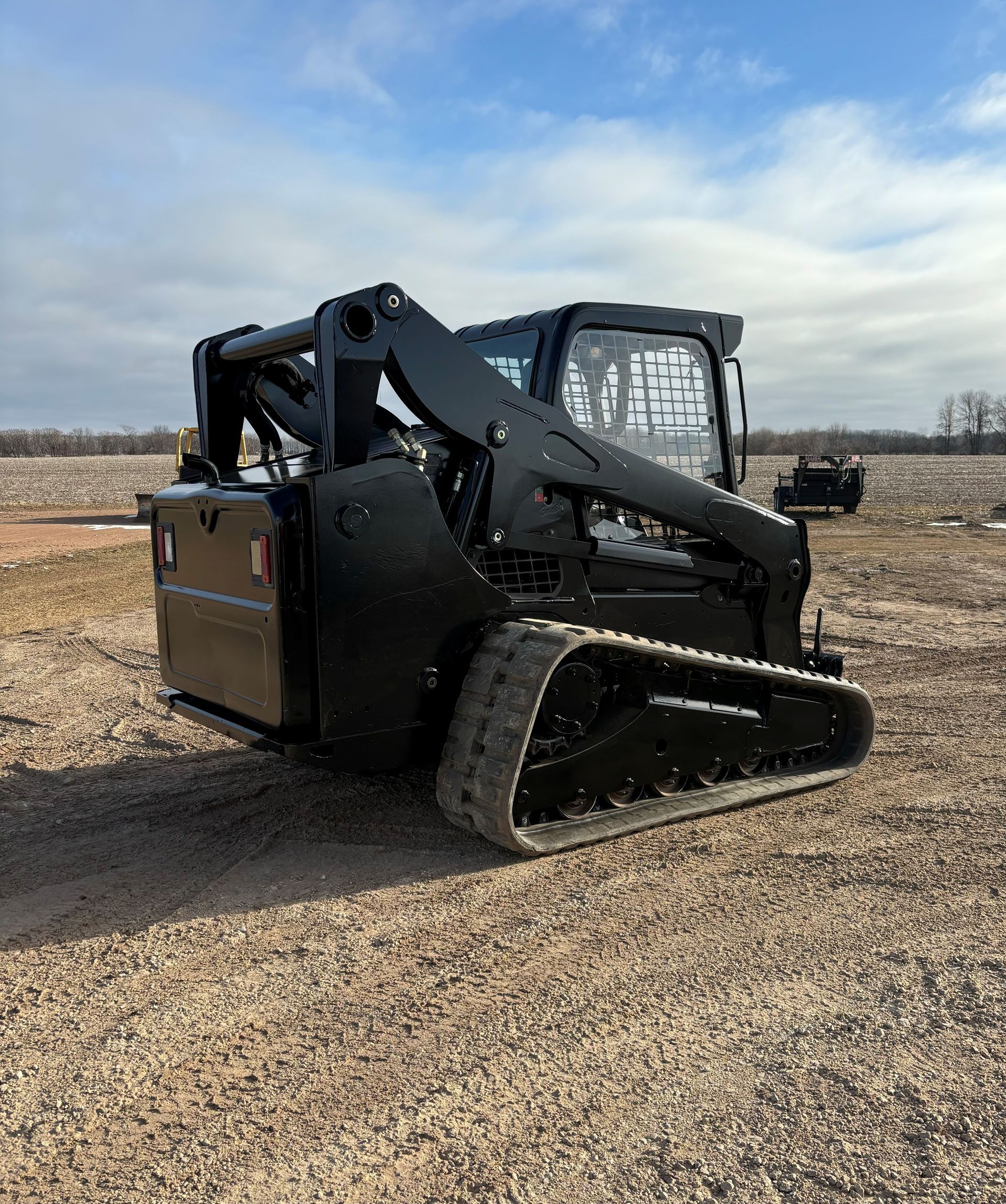 Black track skid steer on gravel field, under cloudy sky.