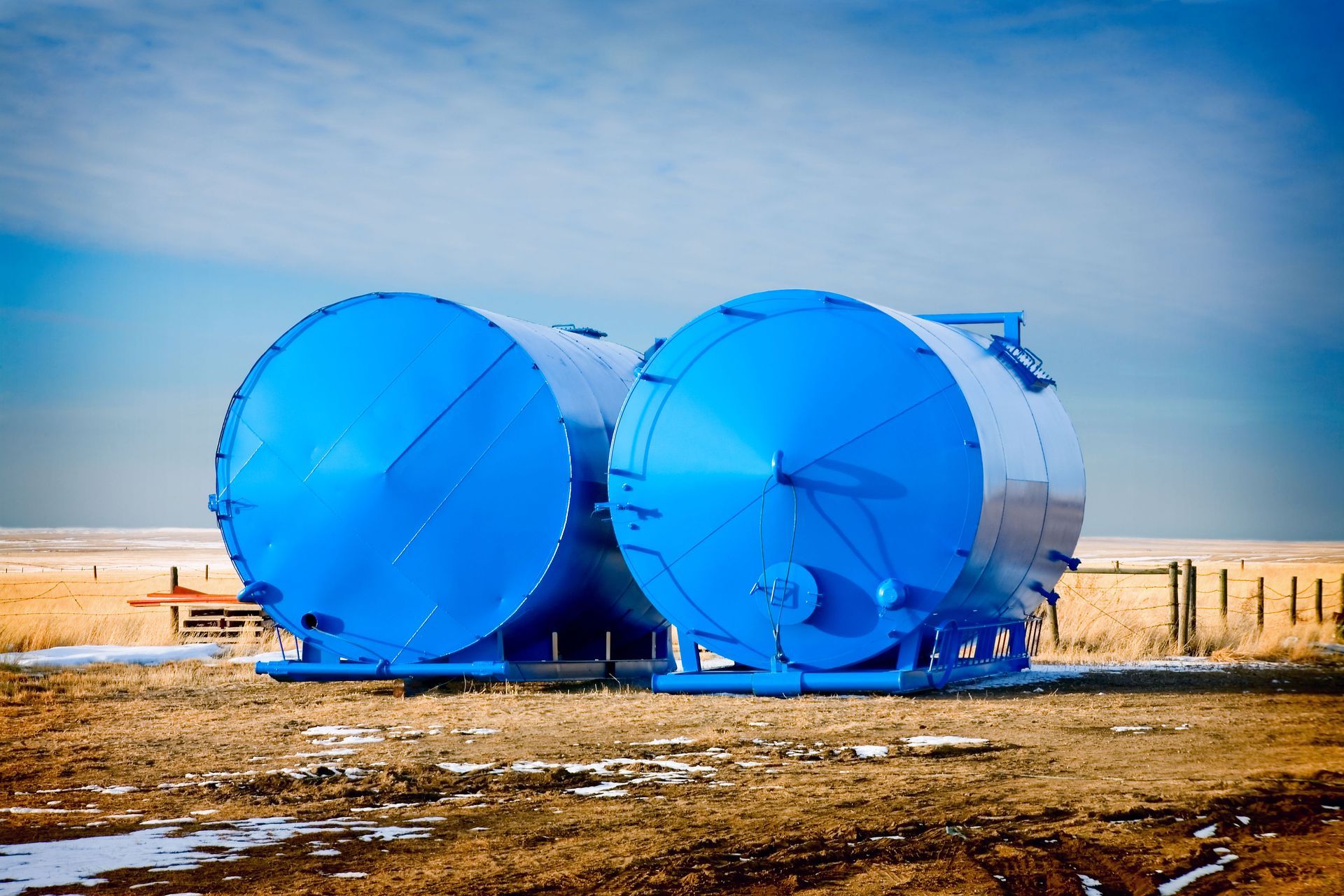Two large blue cylindrical tanks on a brown field, set against a blue sky with sparse vegetation.