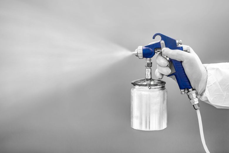 A gloved hand holding a blue spray gun, spraying white paint onto a gray surface.