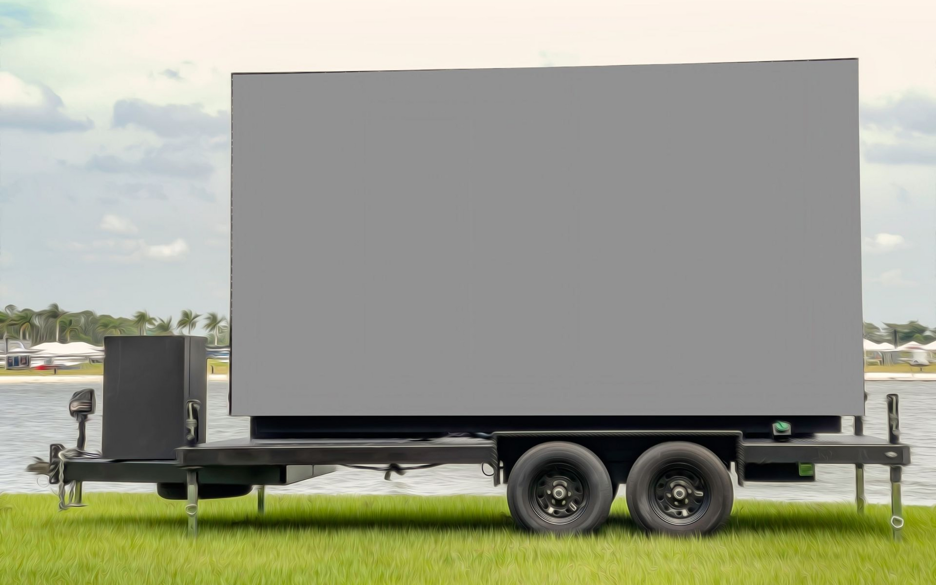 Large, grey, blank LED screen mounted on a black trailer parked on grass near water.