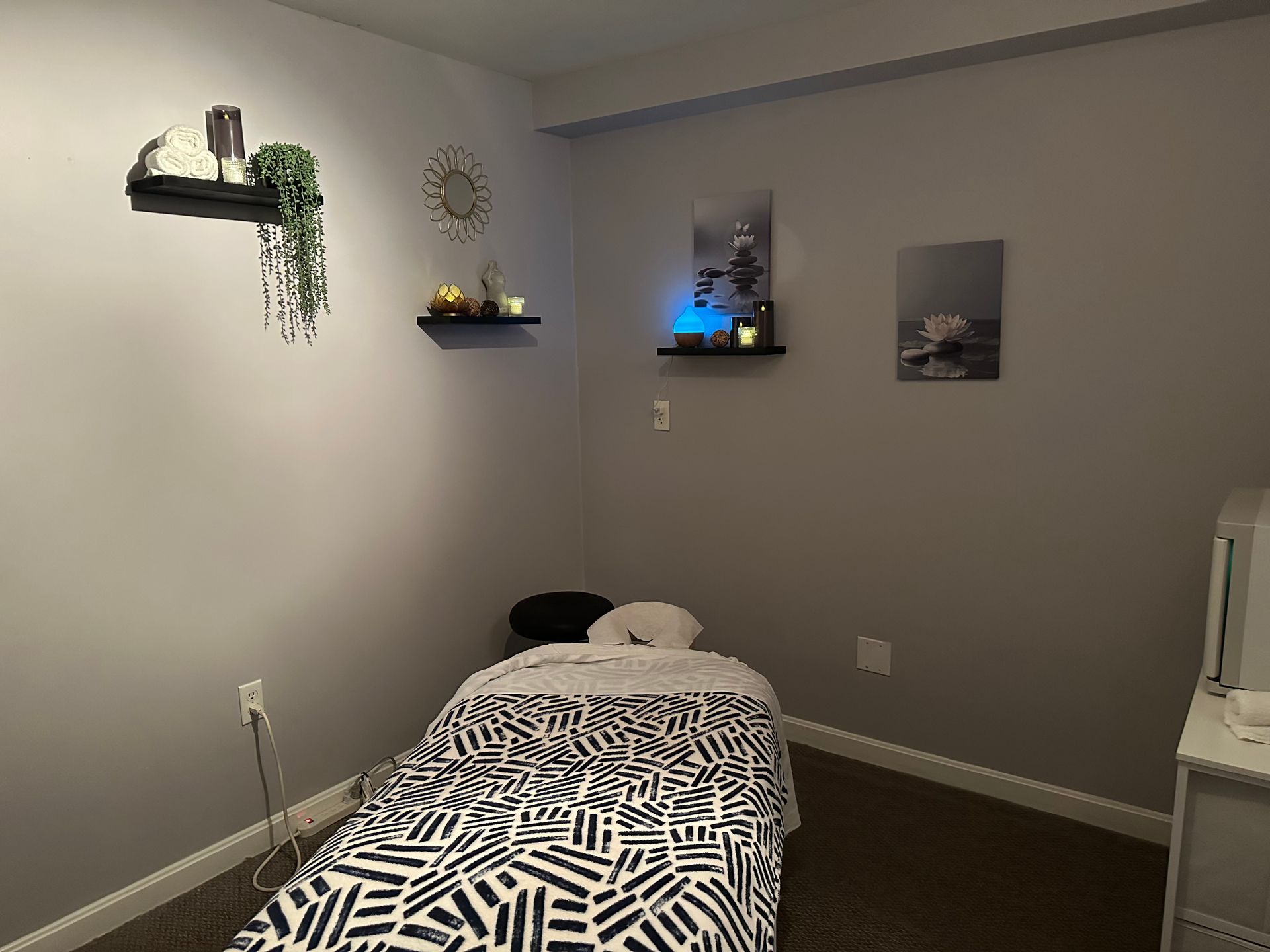 Massage therapy room, gray walls, bed with black and white patterned sheet. Shelves with decor.