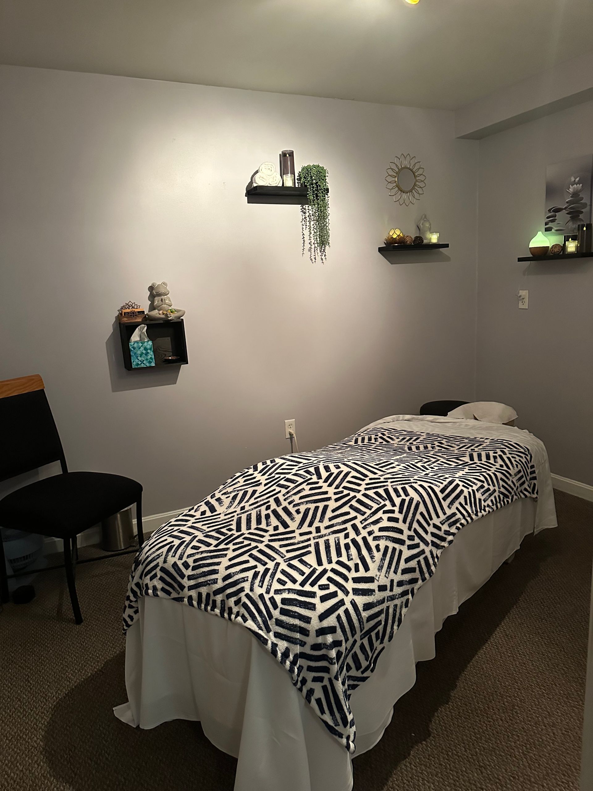 Massage room with a massage table, chair, and shelves with decorative items on a grey wall.