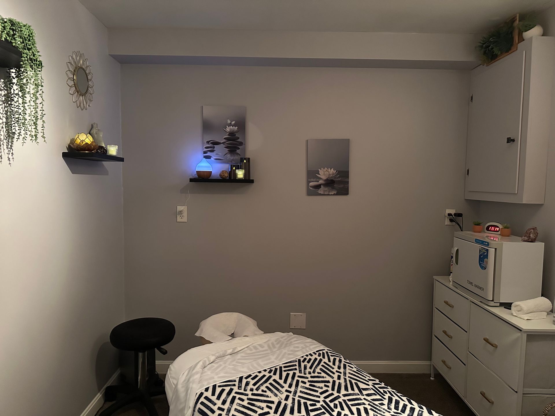 Massage therapy room with bed, stool, dresser, shelves, and gray walls.